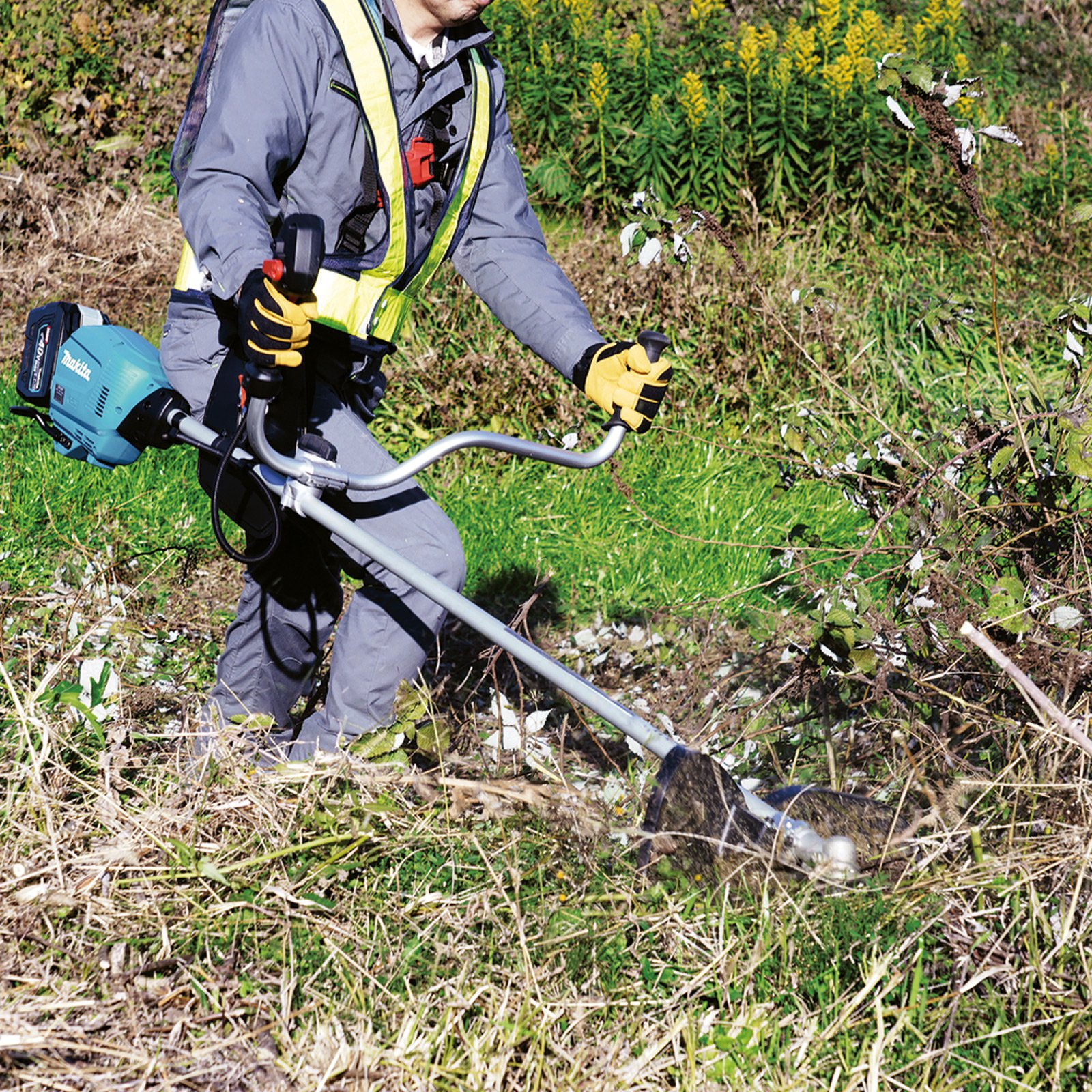 Osoba w ubraniu roboczym używa podkaszarki do koszenia trawy. Narzędzie jest niebiesko-czarne. Żółte kwiaty w tle.