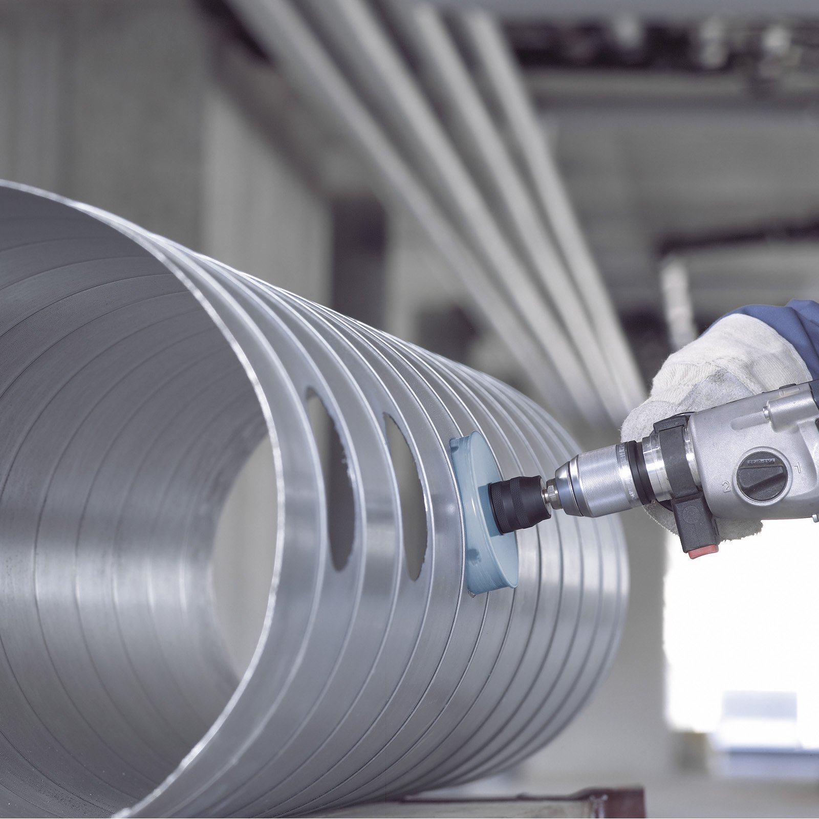 Ein Arbeiter verwendet einen Bohrer mit Schleifaufsatz an einem großen Metallrohr. Die Szene spielt in einer Fabrik.