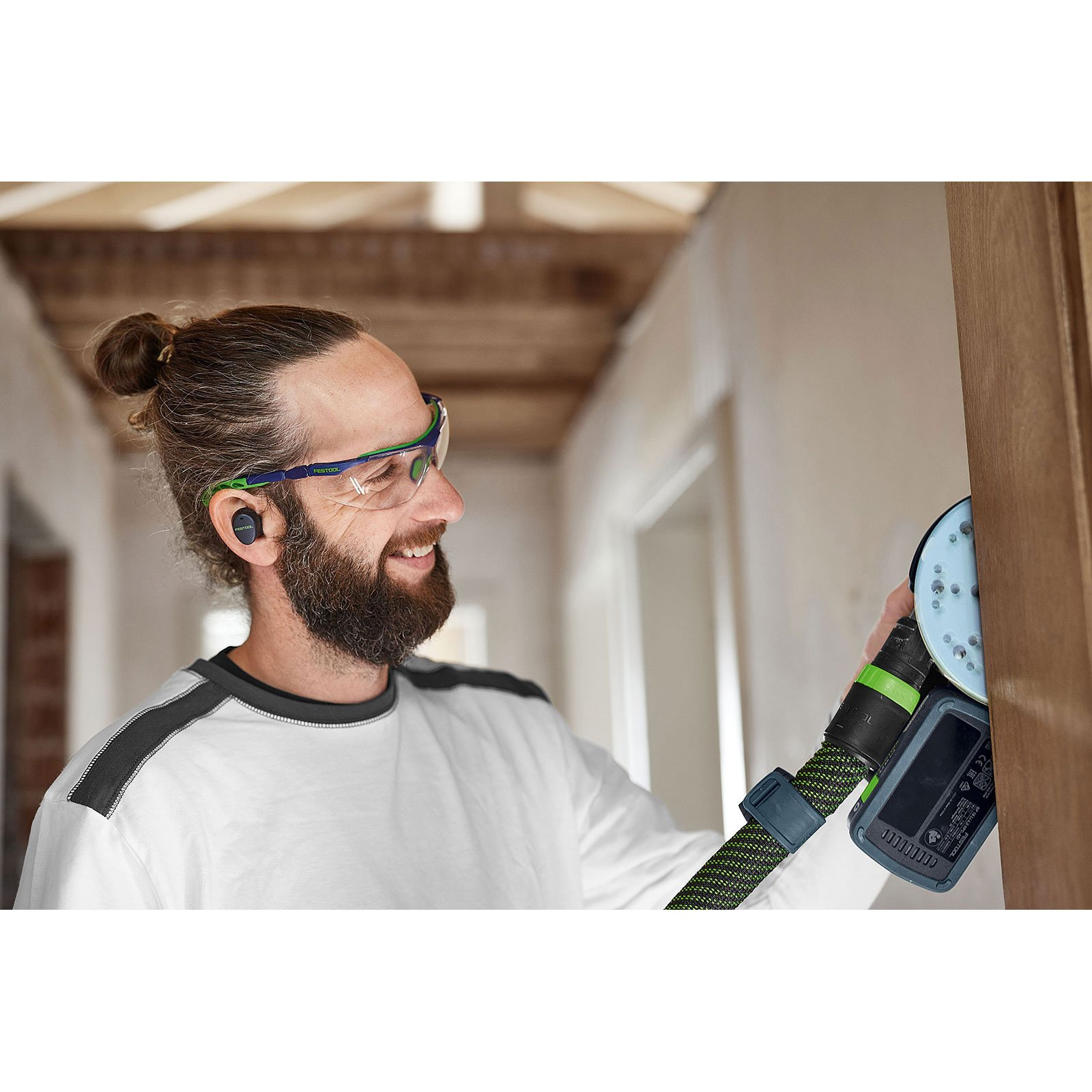 Uomo con barba e chignon con occhiali protettivi e auricolare. Sta carteggiando un telaio di legno con levigatrice.