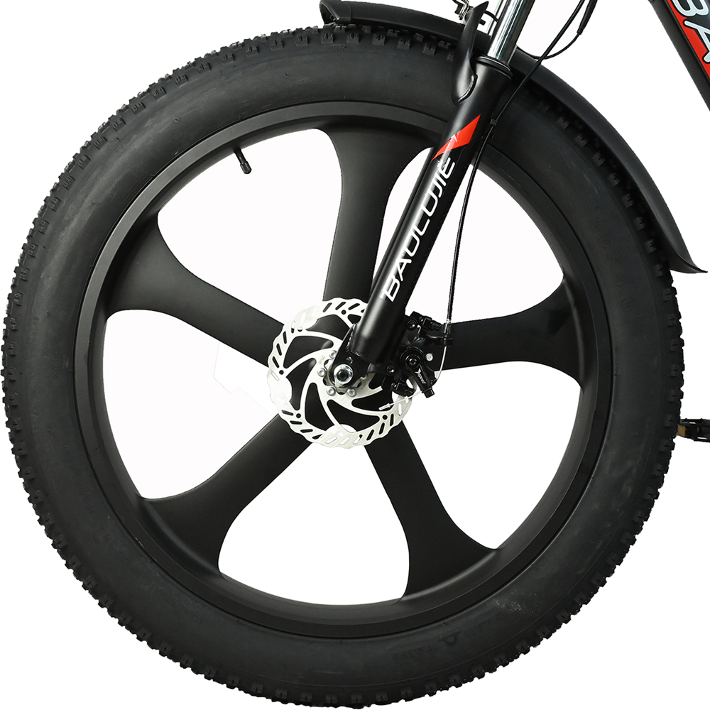 Primer plano de una rueda de bicicleta negra. Tiene un neumático negro, una llanta negra con cinco radios y un freno de disco blanco. La horquilla es negra.