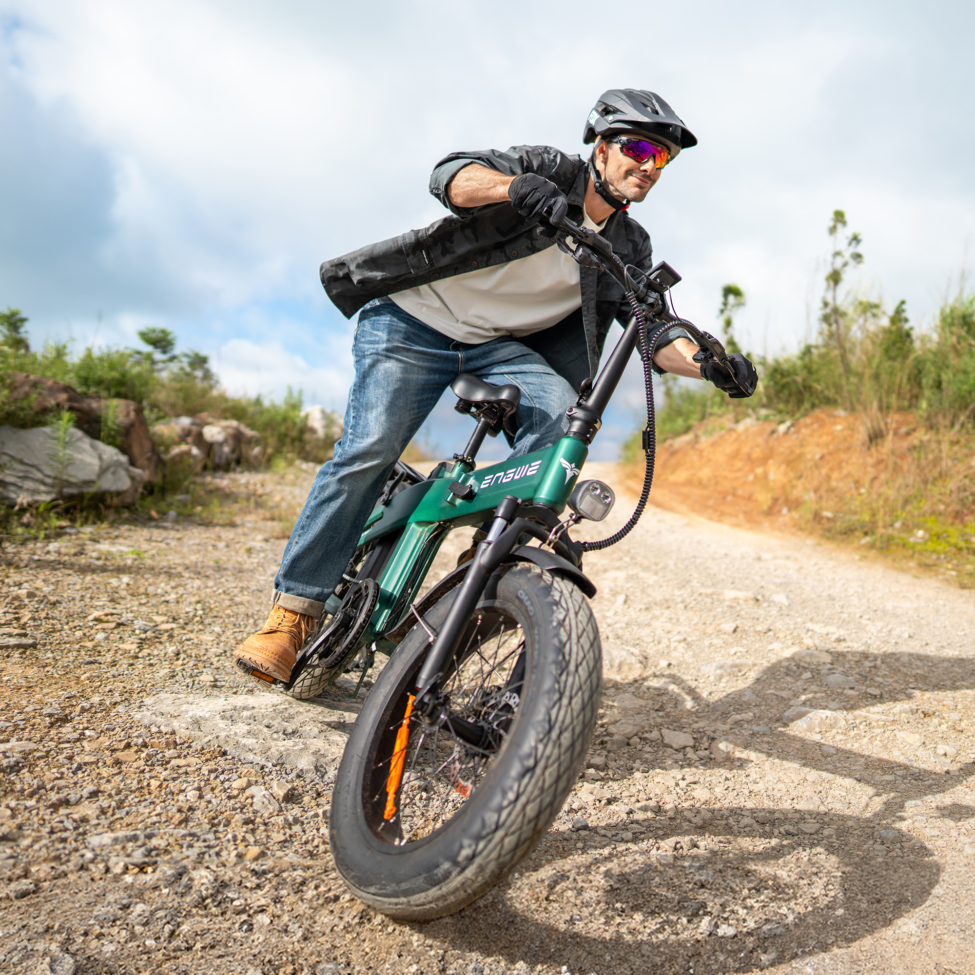 Uomo in sella a una e-bike verde su una strada sterrata. Indossa casco e occhiali da sole. La bici ha pneumatici larghi.