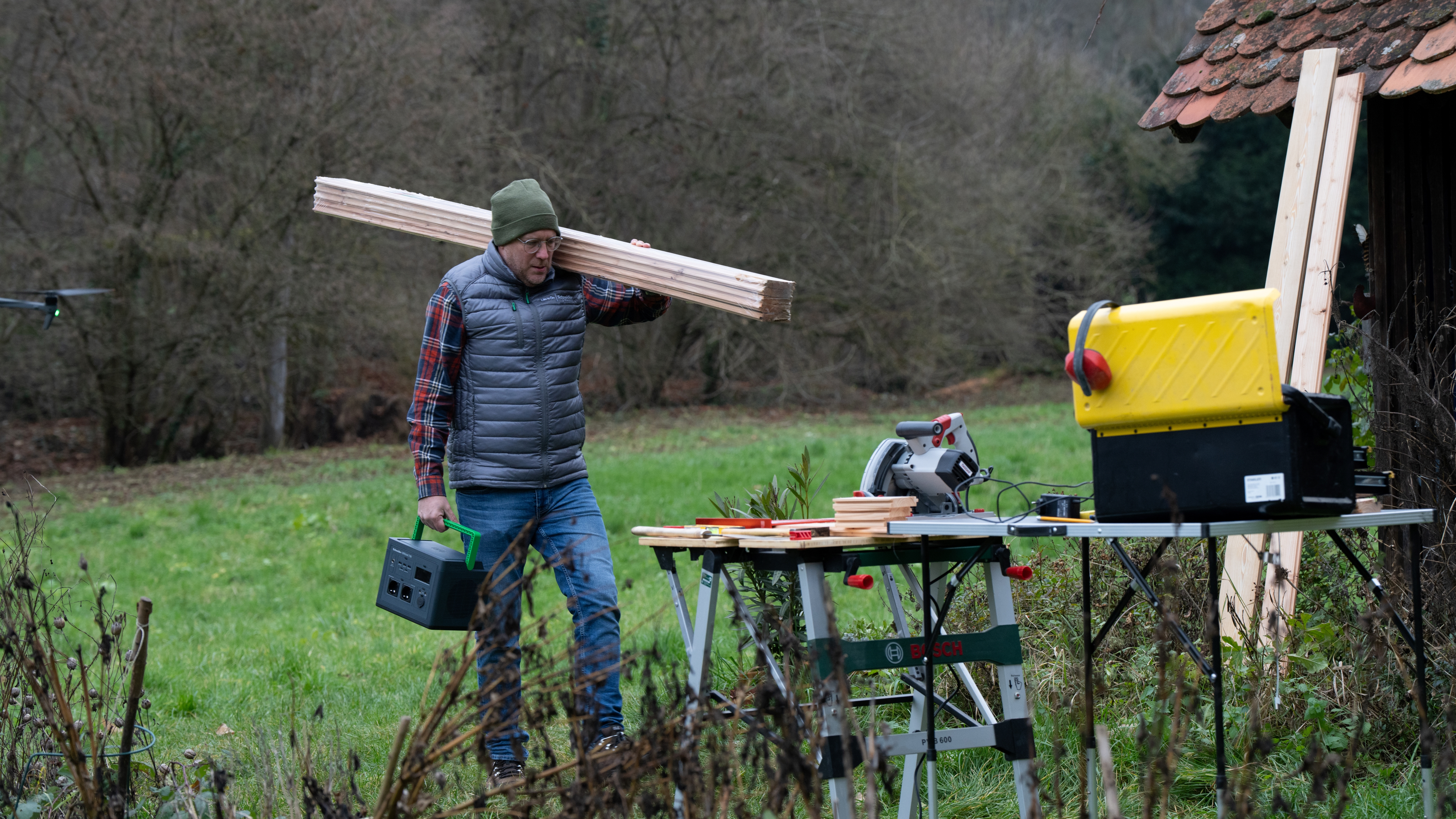 Mężczyzna niesie drewno na ramieniu w pobliżu stołu do piłowania. W tle lata dron. Widoczne są również narzędzia i żółta skrzynka.