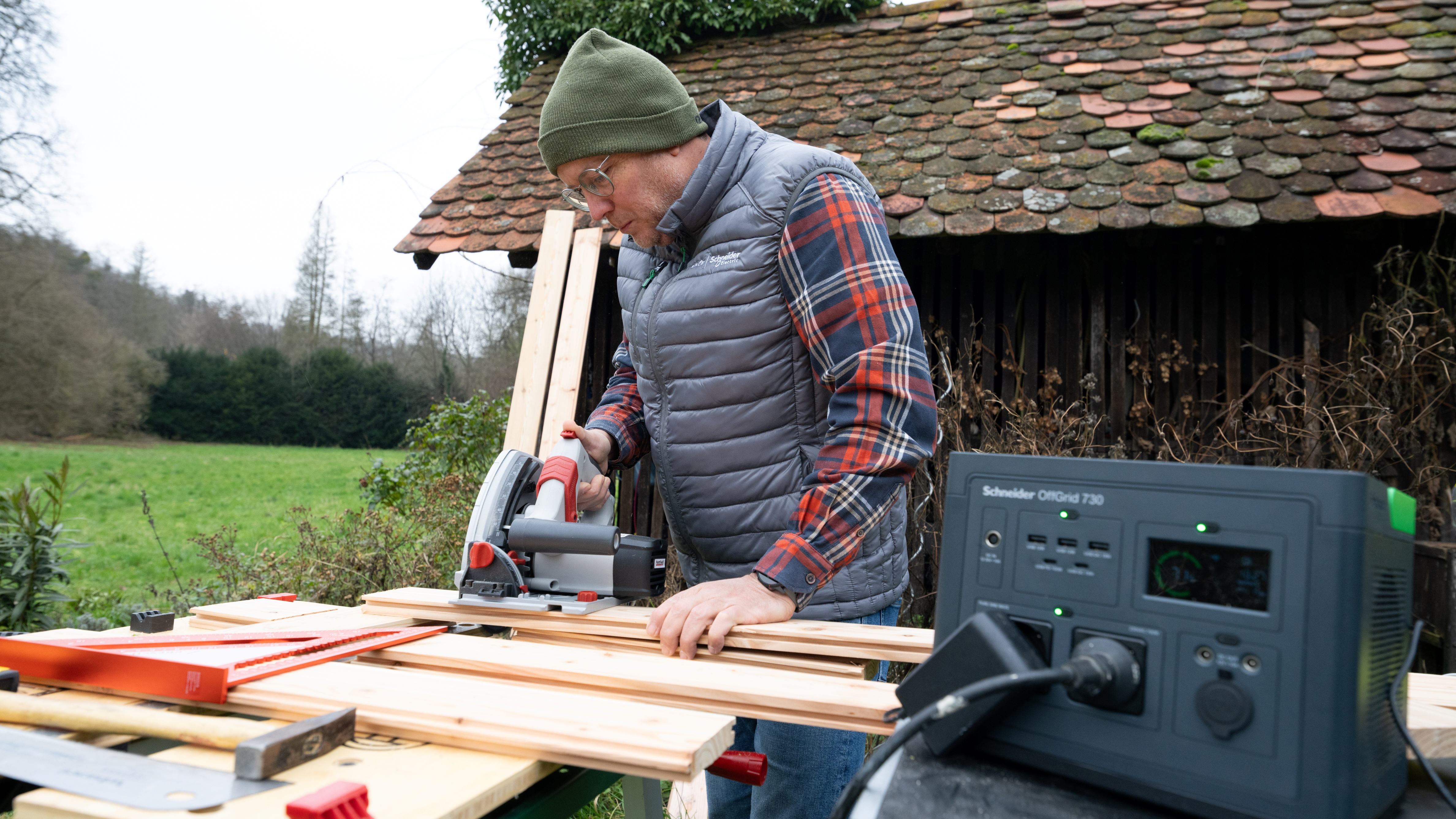 Mężczyzna w zielonej czapce i kamizelce używa piły tarczowej do cięcia drewna. W tle elektrownia, obok starej drewnianej szopy.