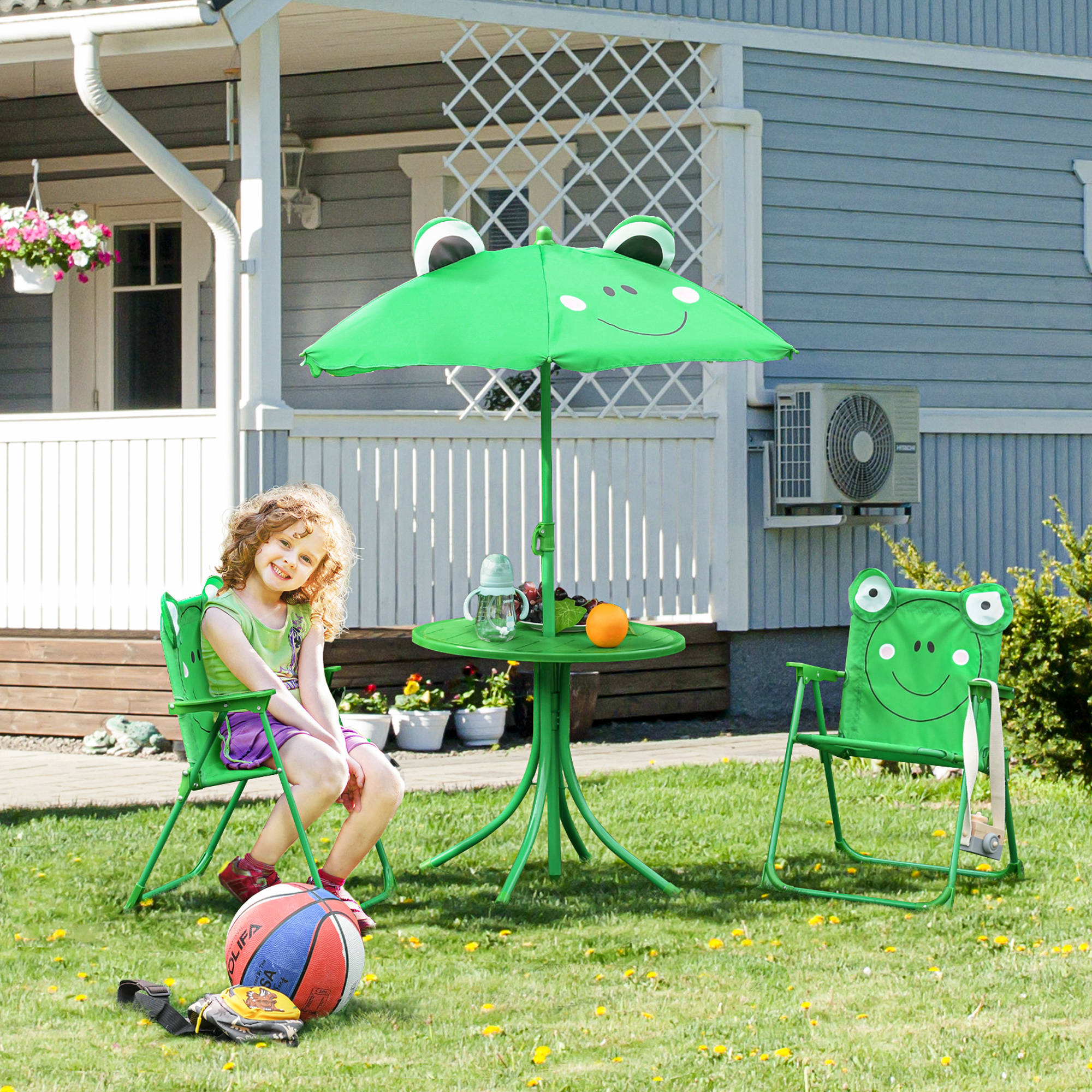 Una bambina seduta su una sedia verde, vicino a un tavolo e a un ombrellone a tema rana. Pallone da basket e guanto sull'erba.