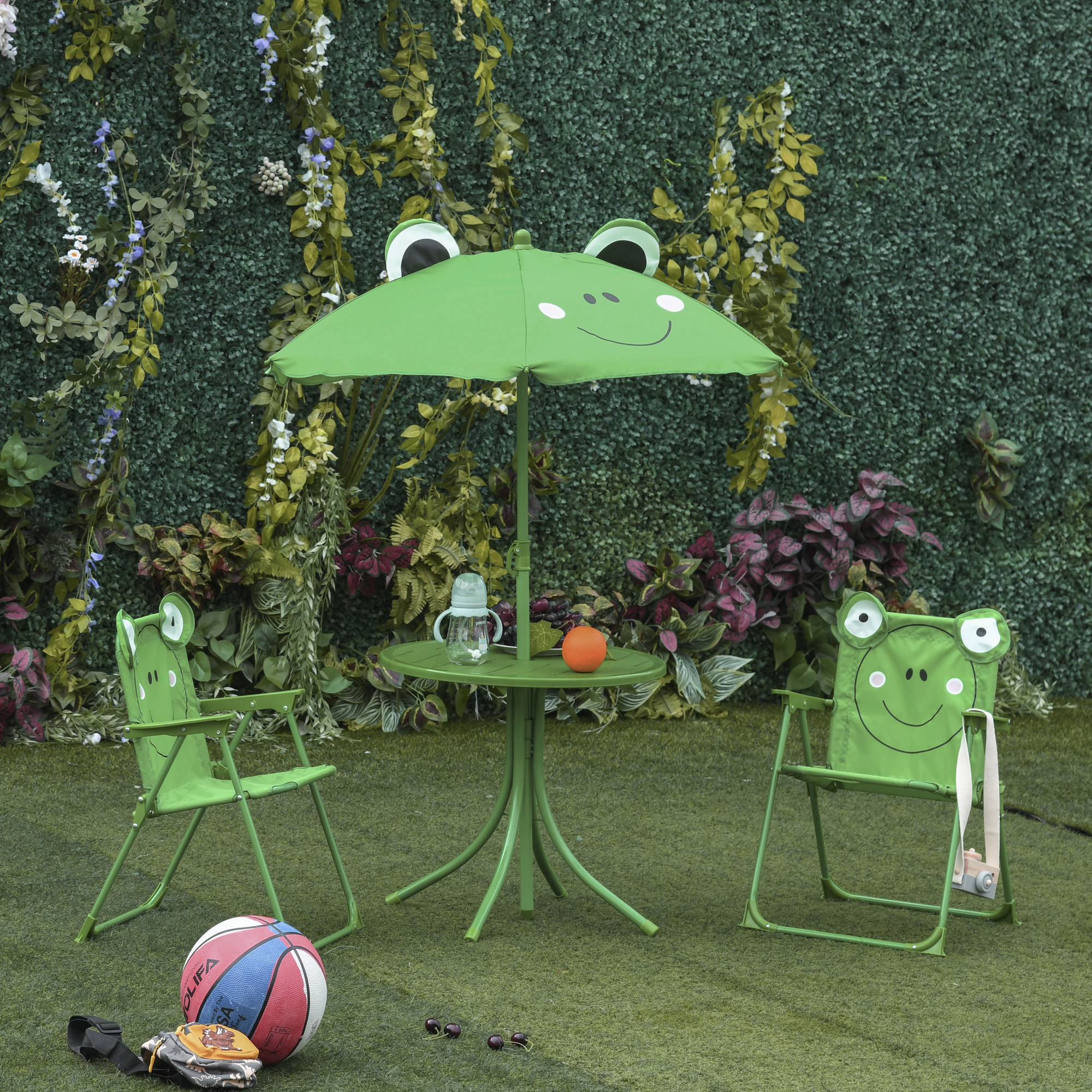 Un set da patio a tema rana verde con ombrellone, tavolo, due sedie e una palla sull'erba verde. Lo sfondo ha vegetazione.