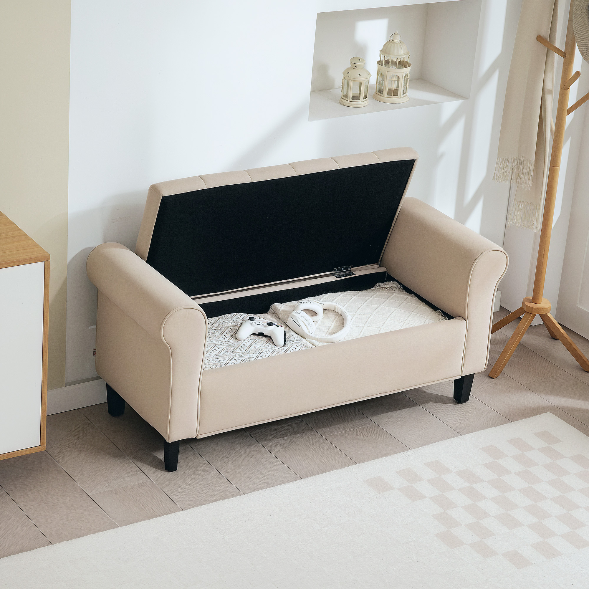 Panca contenitore beige con coperchio aperto, che rivela coperta, controller e cuffie. Un attaccapanni sullo sfondo.