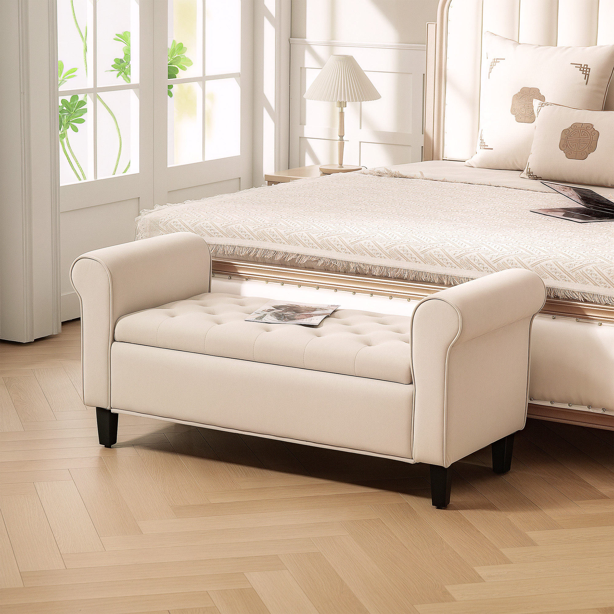 Scena di camera da letto con panca e letto color crema. Finestra con piante a sinistra. Lampada su tavolo e cuscini sul letto.