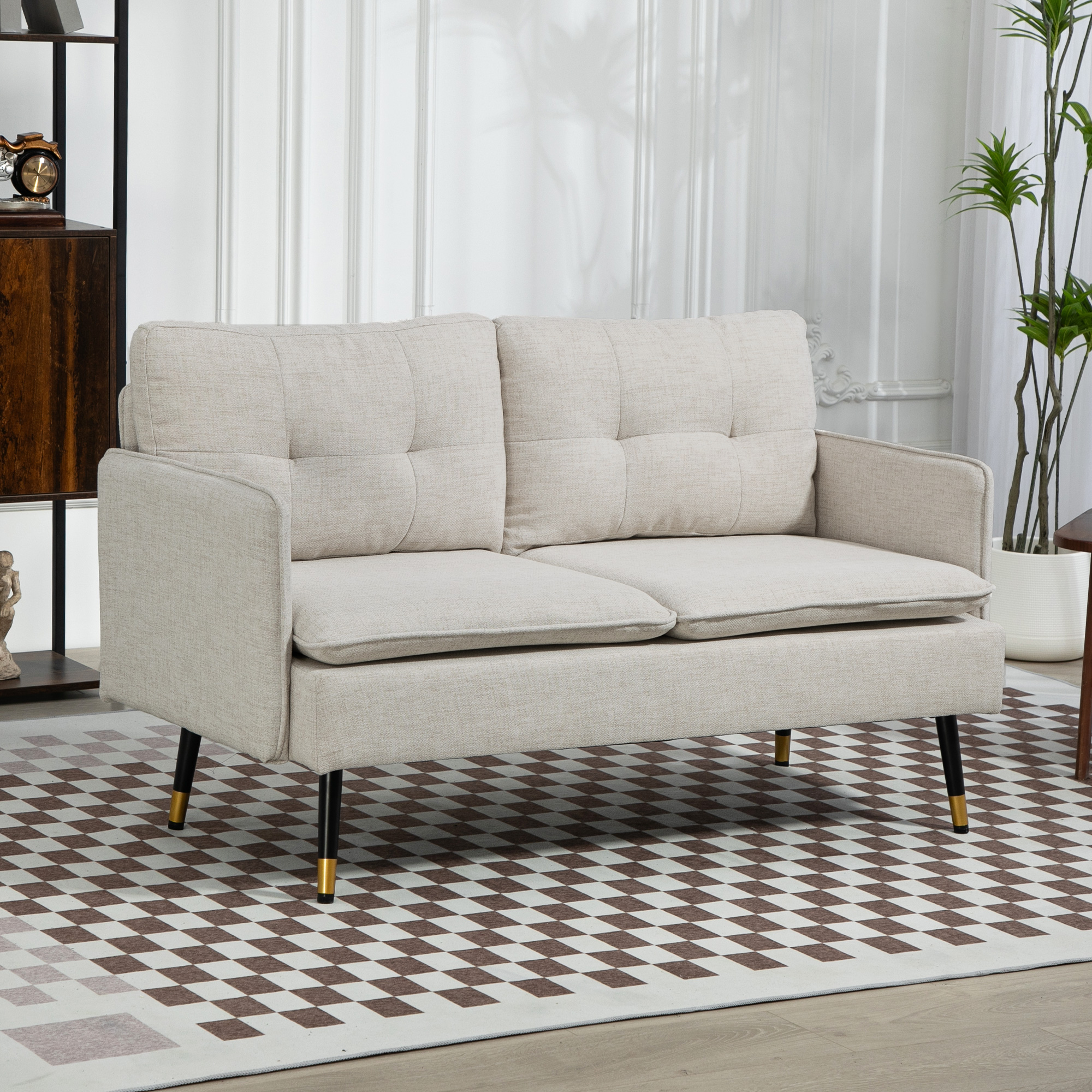 Divano beige a due posti con gambe nere e dorate su un tappeto a scacchi, vicino a una pianta e un mobile.
