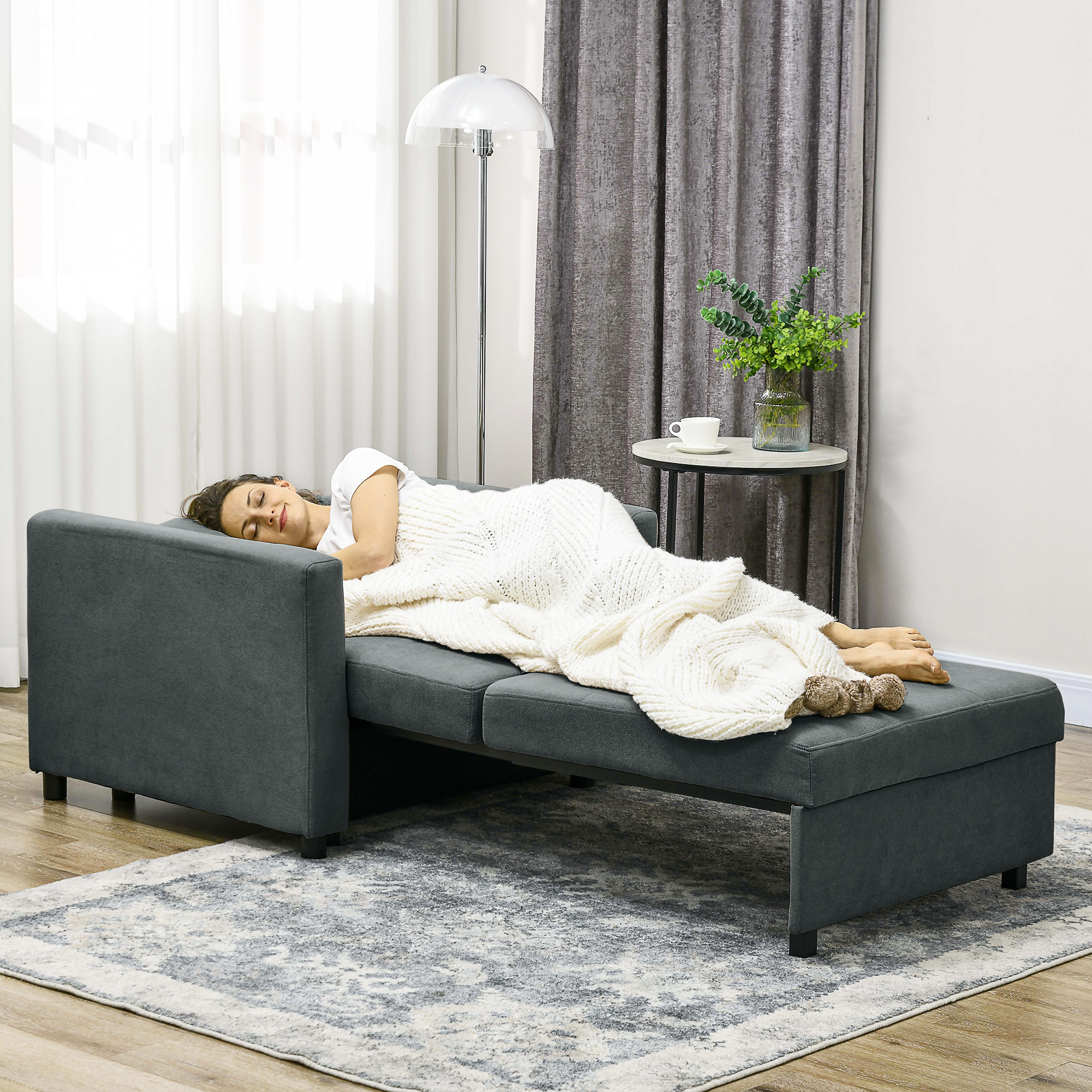 Una donna dorme su un divano grigio, coperta da una coperta bianca. Sono visibili una lampada da terra, tende e un tavolino.