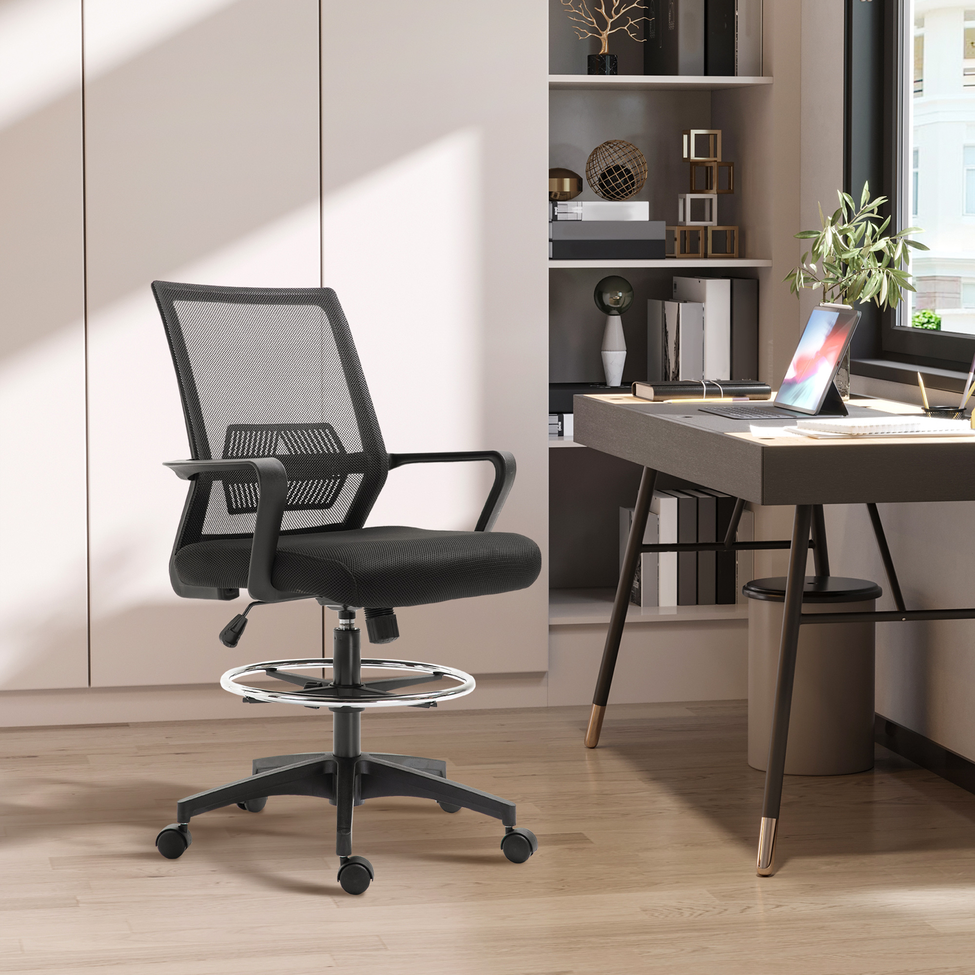 Sedia da ufficio in rete nera. Scrivania con laptop e pianta sullo sfondo. Pavimento in legno e parete bianca.