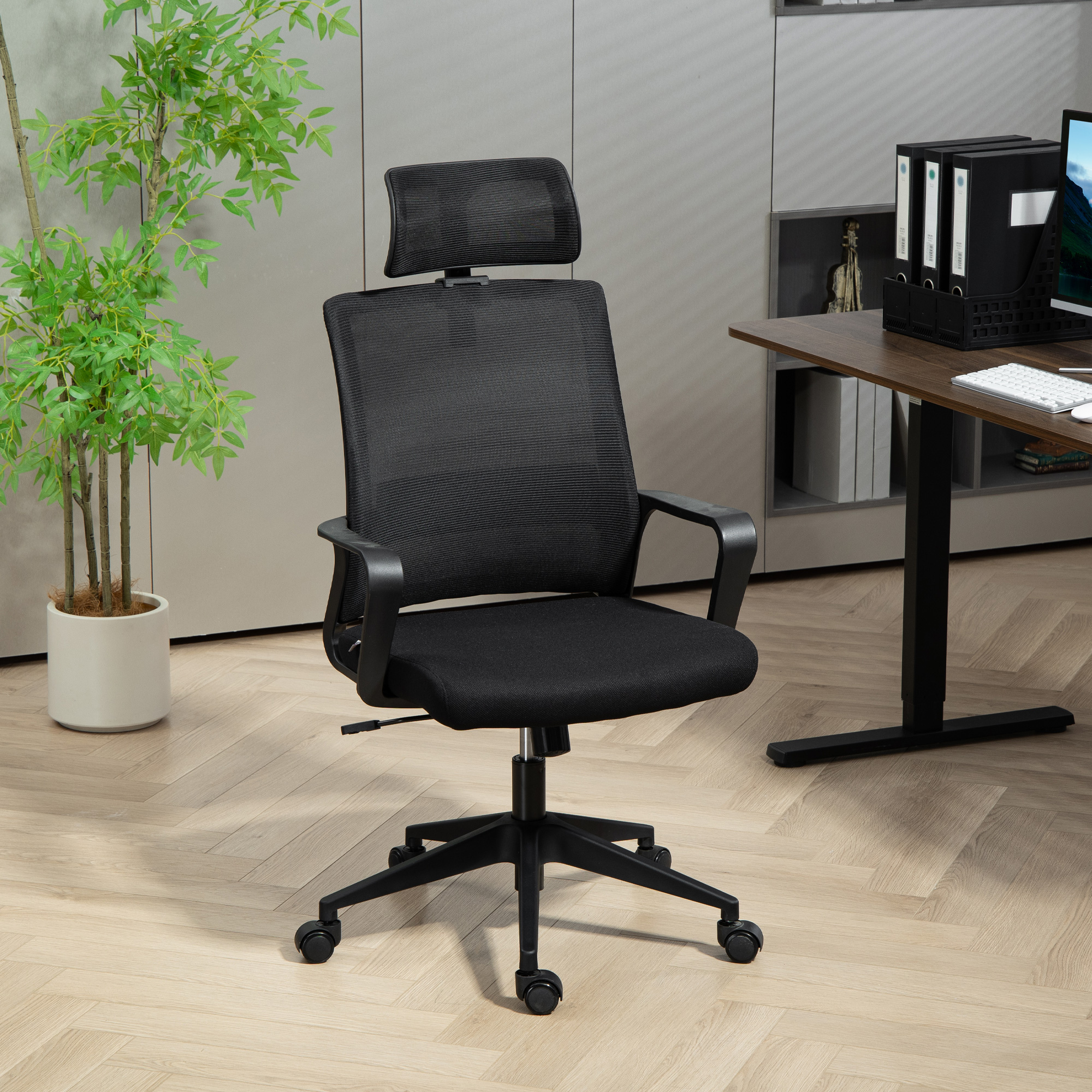 Una sedia da ufficio nera in rete con poggiatesta, in un ambiente ufficio. Sono visibili anche una pianta, una scrivania e un computer.