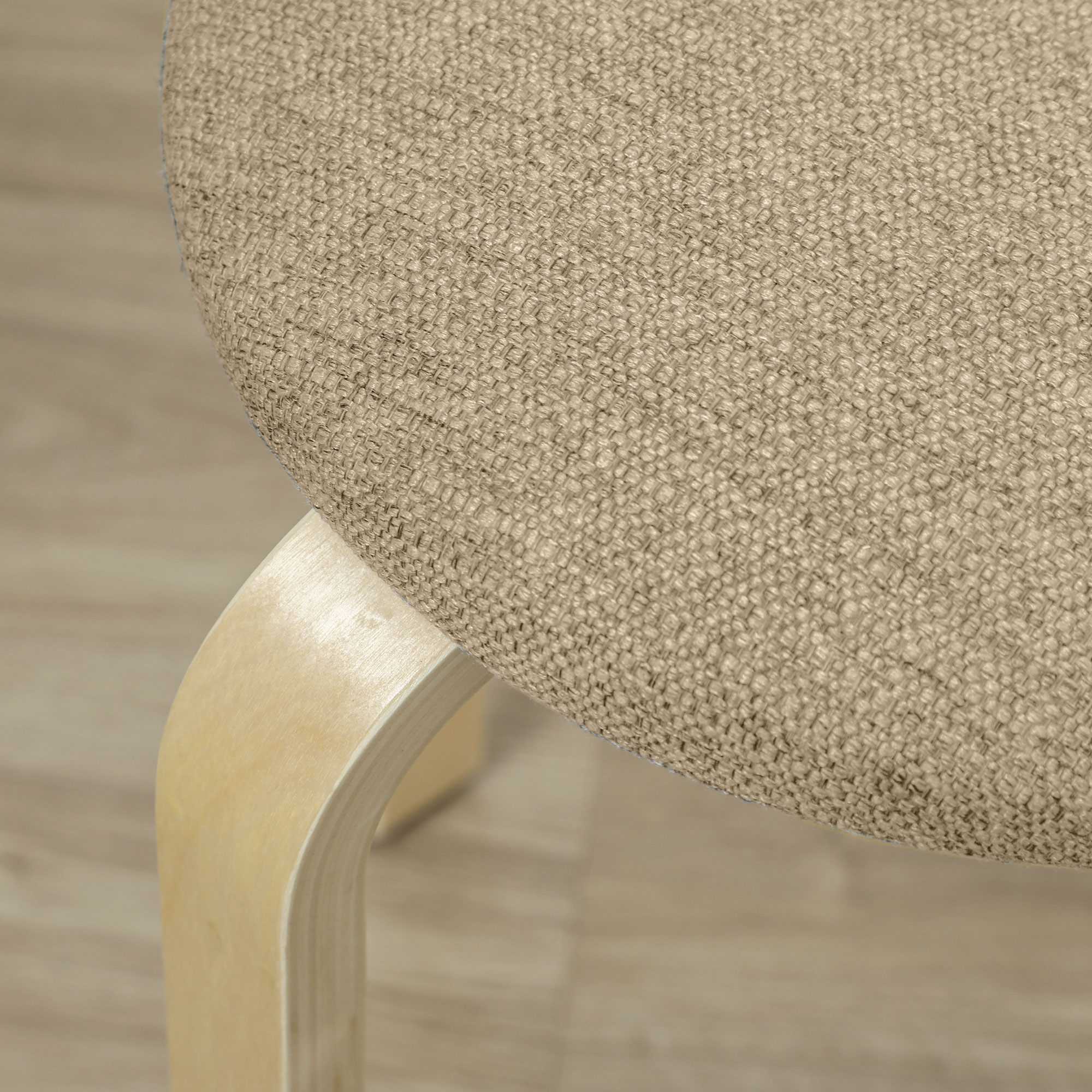 Primo piano di un sedile in tessuto beige e una gamba in legno di uno sgabello. Lo sfondo è un pavimento in legno chiaro.