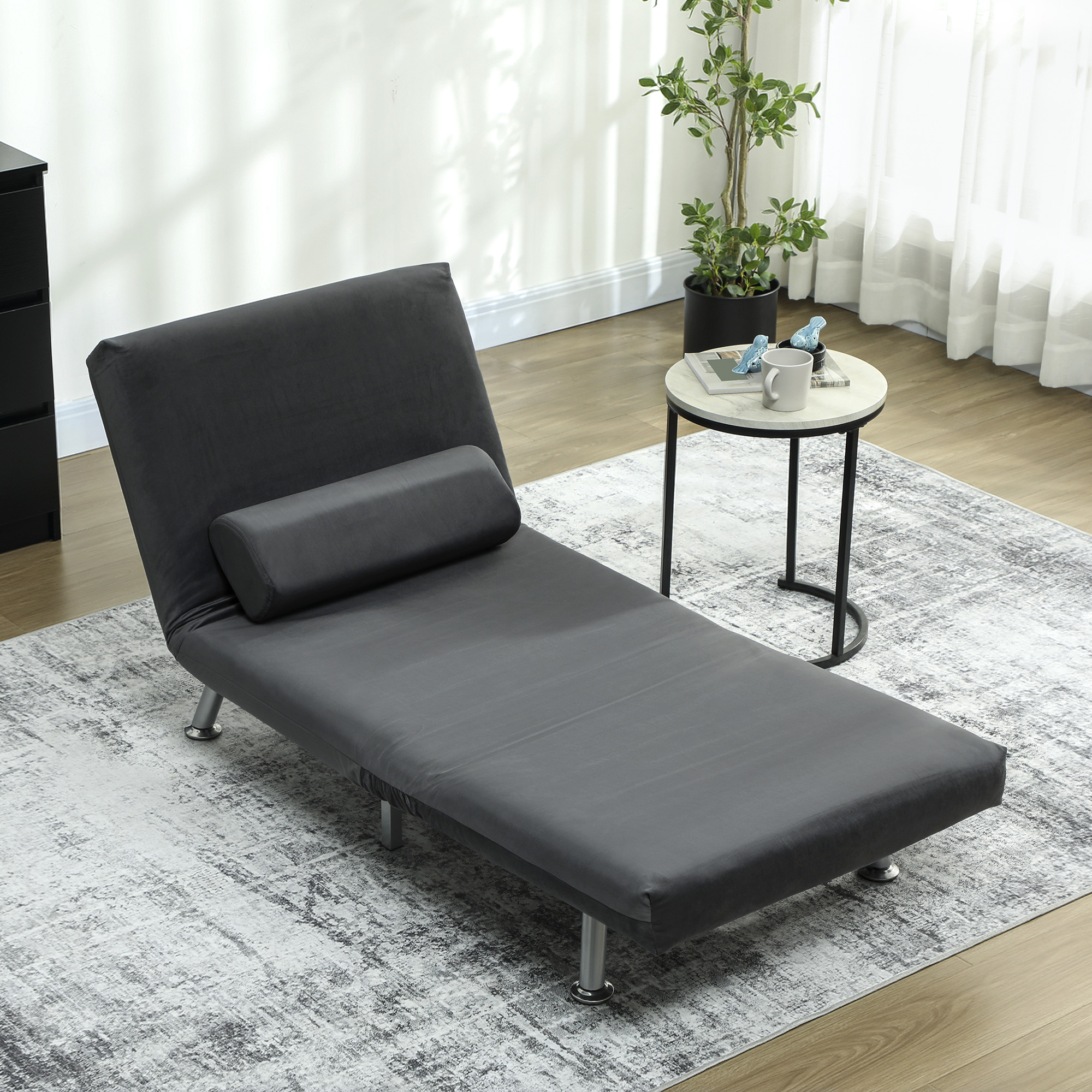 Una chaise longue grigia con un cuscino a rullo è su un tappeto fantasia. Un tavolino e una finestra con tende sono sullo sfondo.