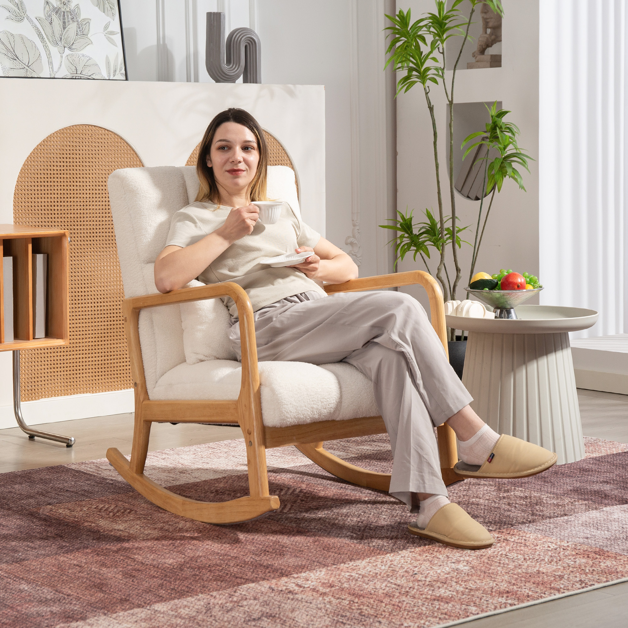 Donna in sedia a dondolo con tazza e piattino. Tavolo con frutta e tappeto visibili.