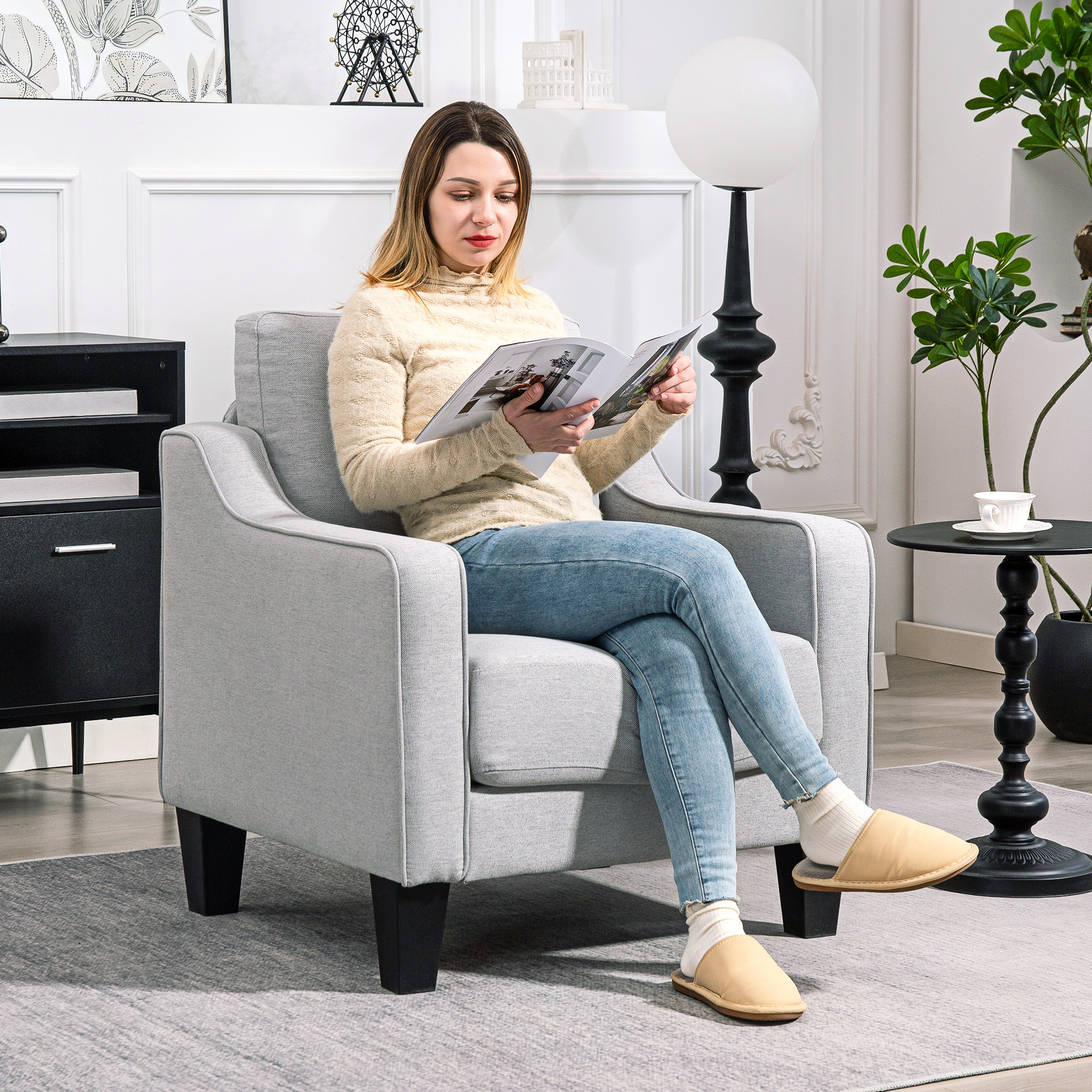 Una donna con un maglione beige e jeans legge una rivista seduta su una poltrona grigia. Una lampada e un tavolo sono vicino a lei.