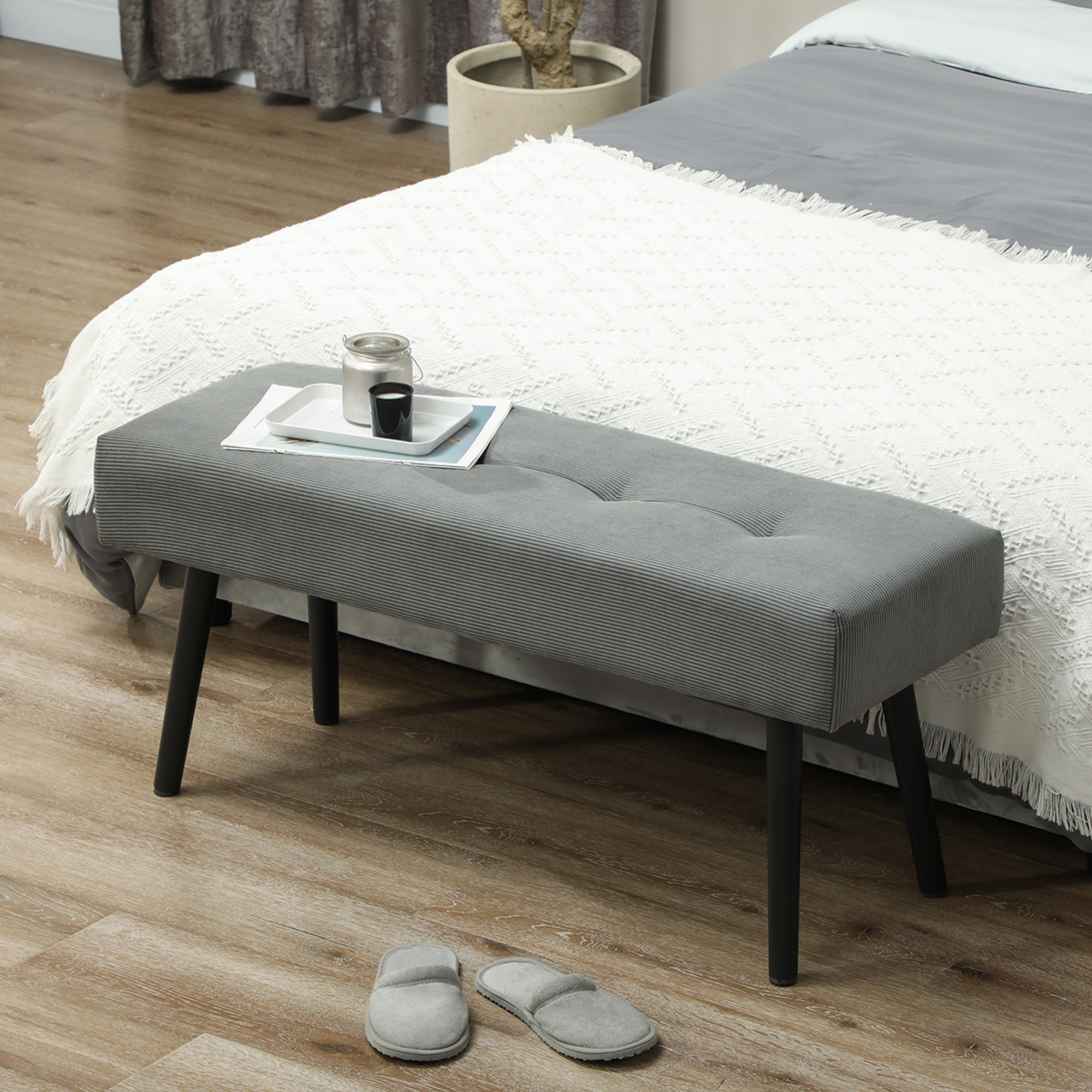 Una panca grigia con gambe nere, un letto con una coperta bianca e pantofole su un pavimento in legno. Un vassoio con oggetti è sulla panca.