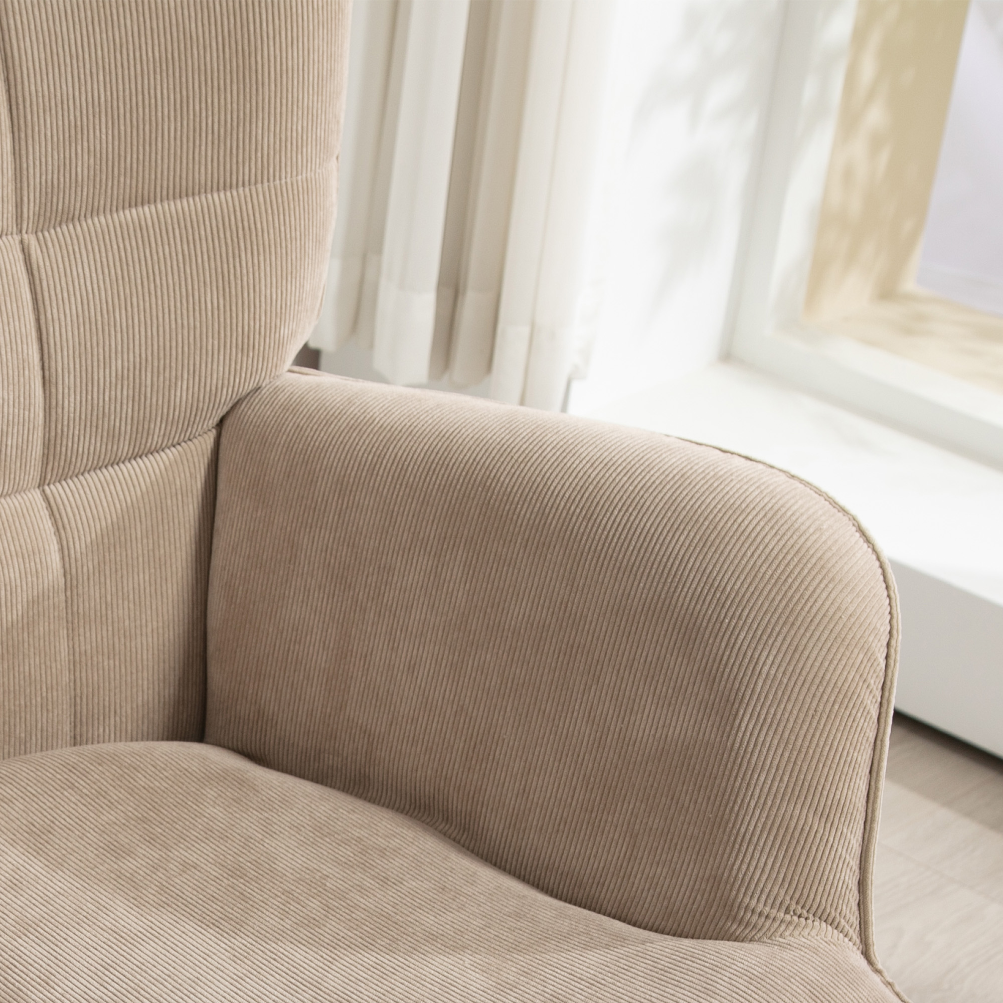 Primo piano di una poltrona in velluto a coste beige. Dettagli: bracciolo, schienale e seduta. Sullo sfondo una finestra con tende trasparenti.