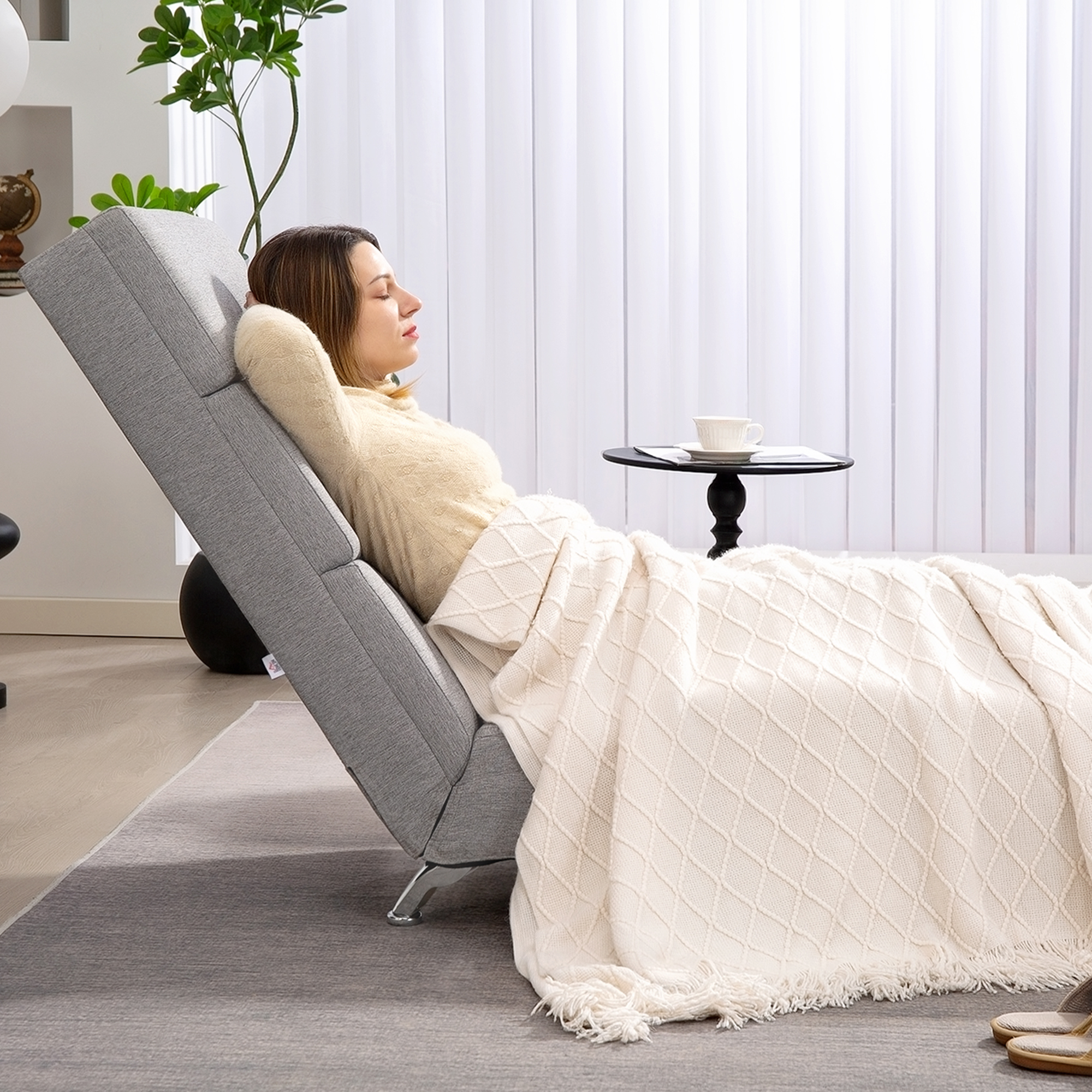 Una donna si riposa su una poltrona grigia, coperta da una coperta color crema. Un tavolino ha una tazza.