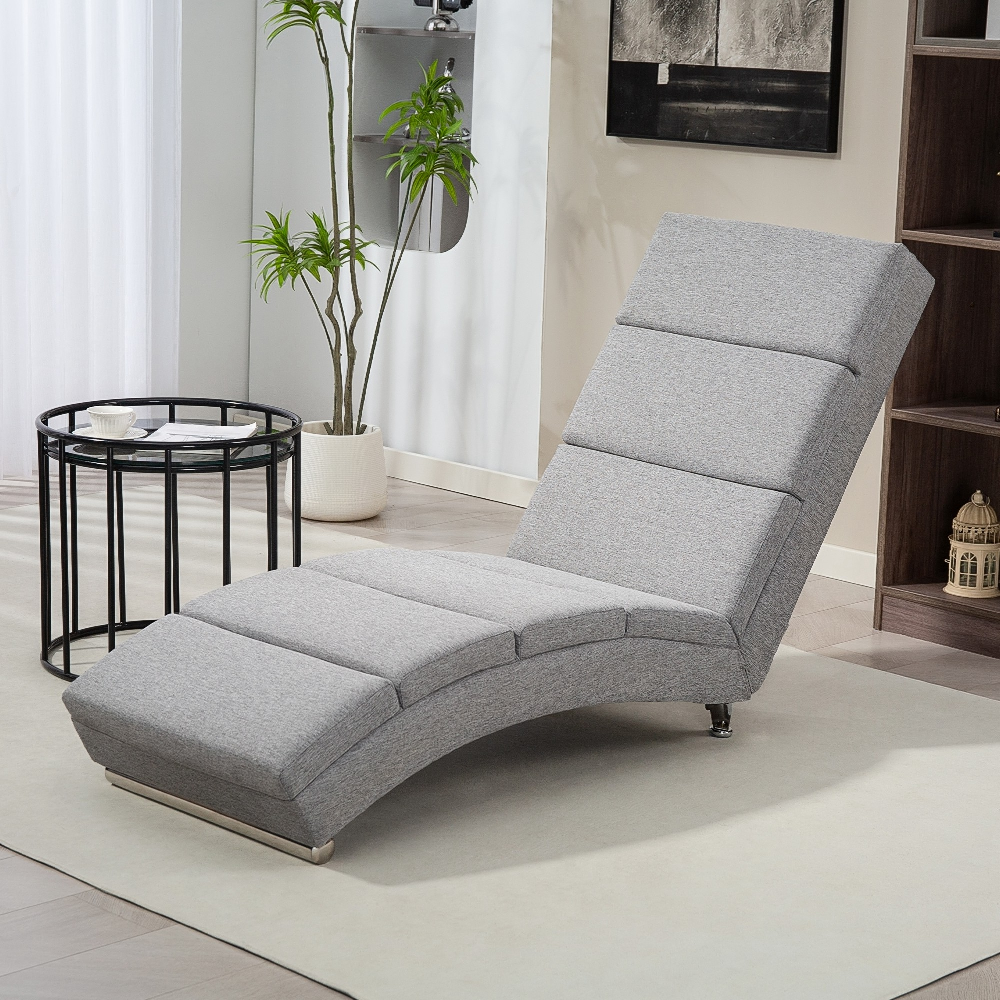 Una chaise longue grigia è su un tappeto bianco. Un tavolino con una tazza e un vaso di piante sono visibili.