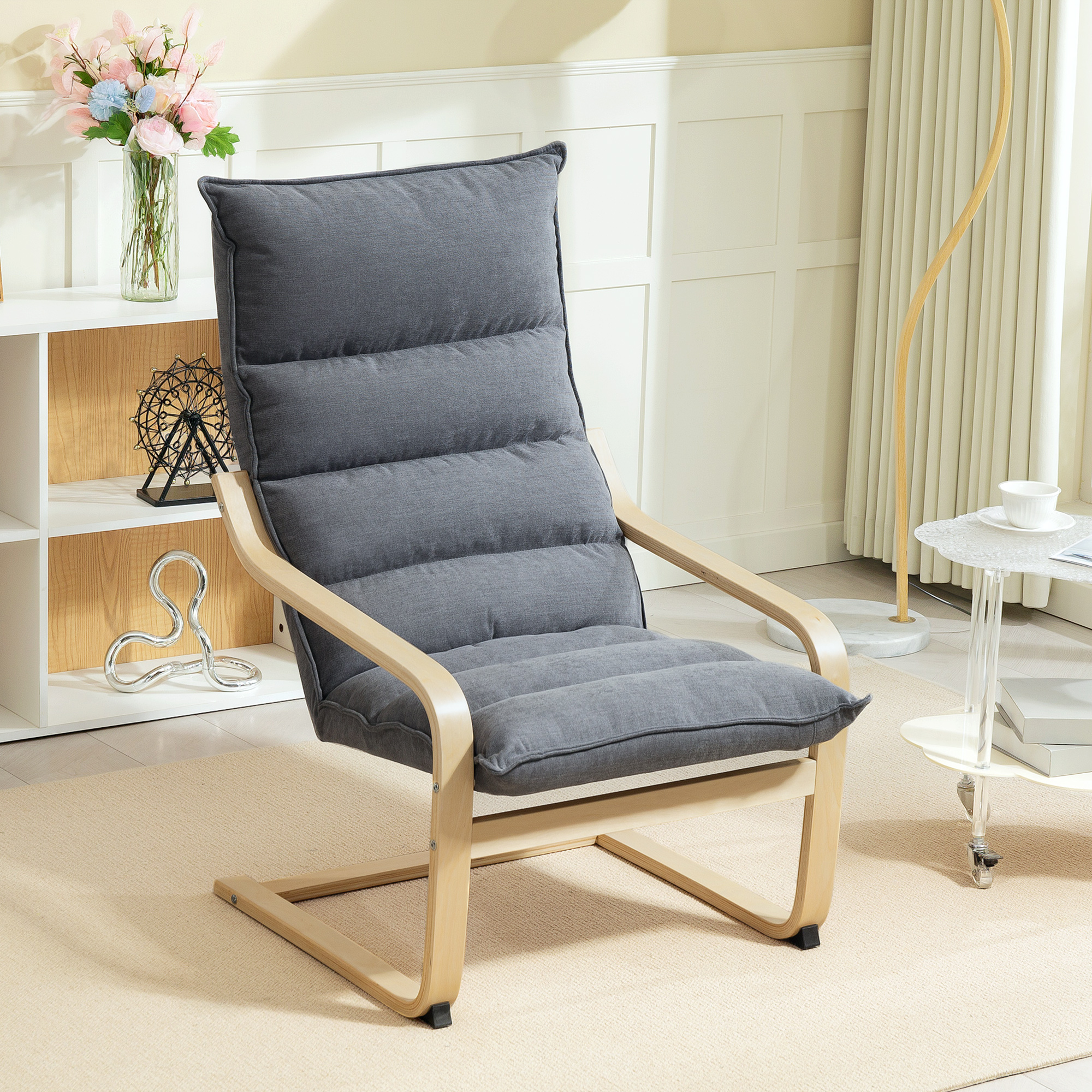 Una sedia grigia imbottita con struttura in legno è su un tappeto beige. Un vaso di fiori e un tavolino sono vicini.