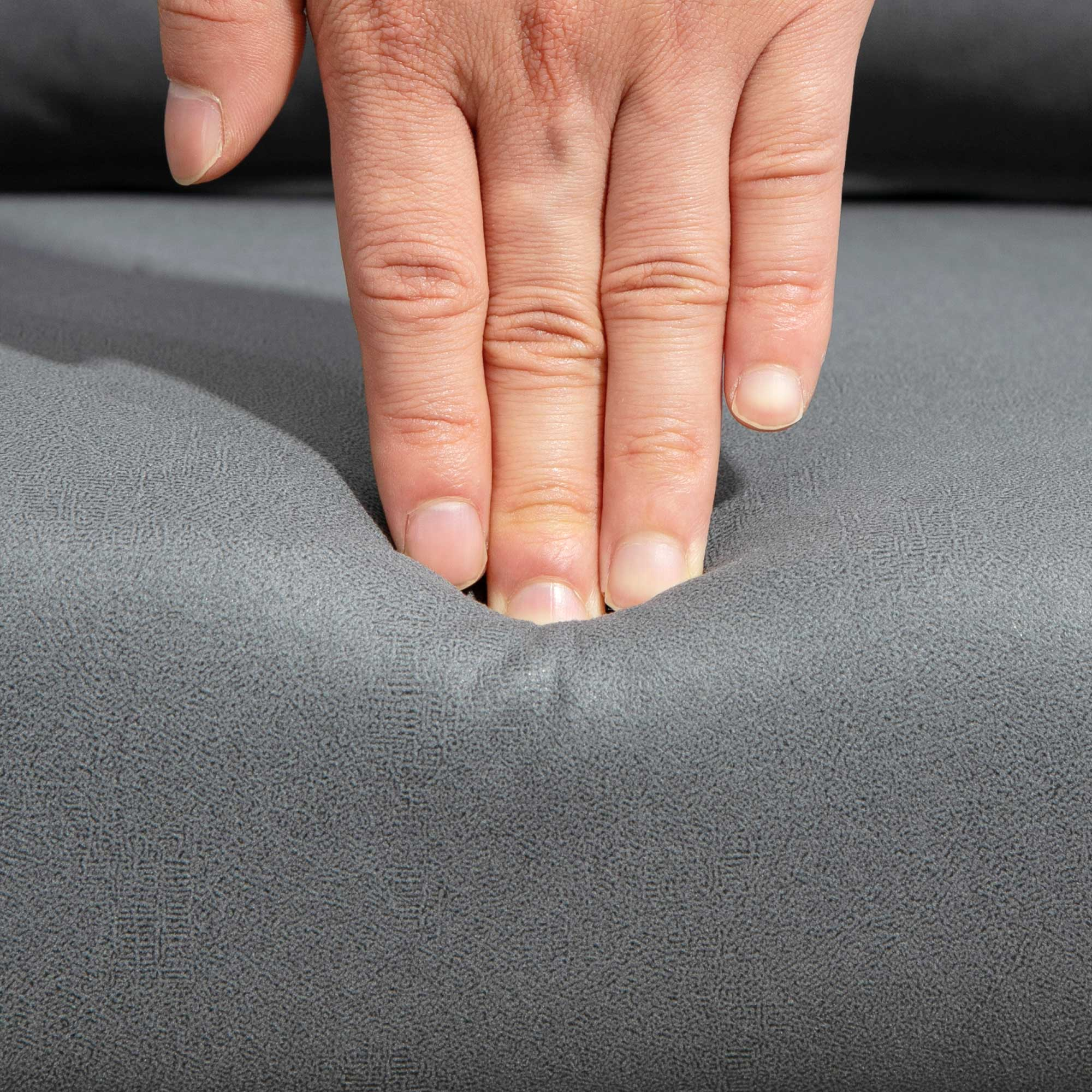 Una mano preme su un tessuto grigio, creando un'indentazione. Le dita e le unghie sono visibili. Lo sfondo è sfocato.