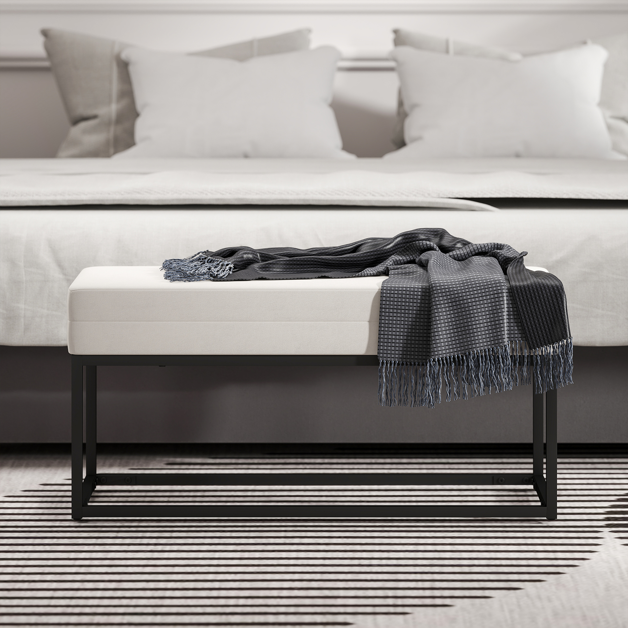 Scena di camera da letto con panca e coperta davanti a un letto. La panca ha una struttura nera e un cuscino chiaro. Una coperta grigia è sulla panca.