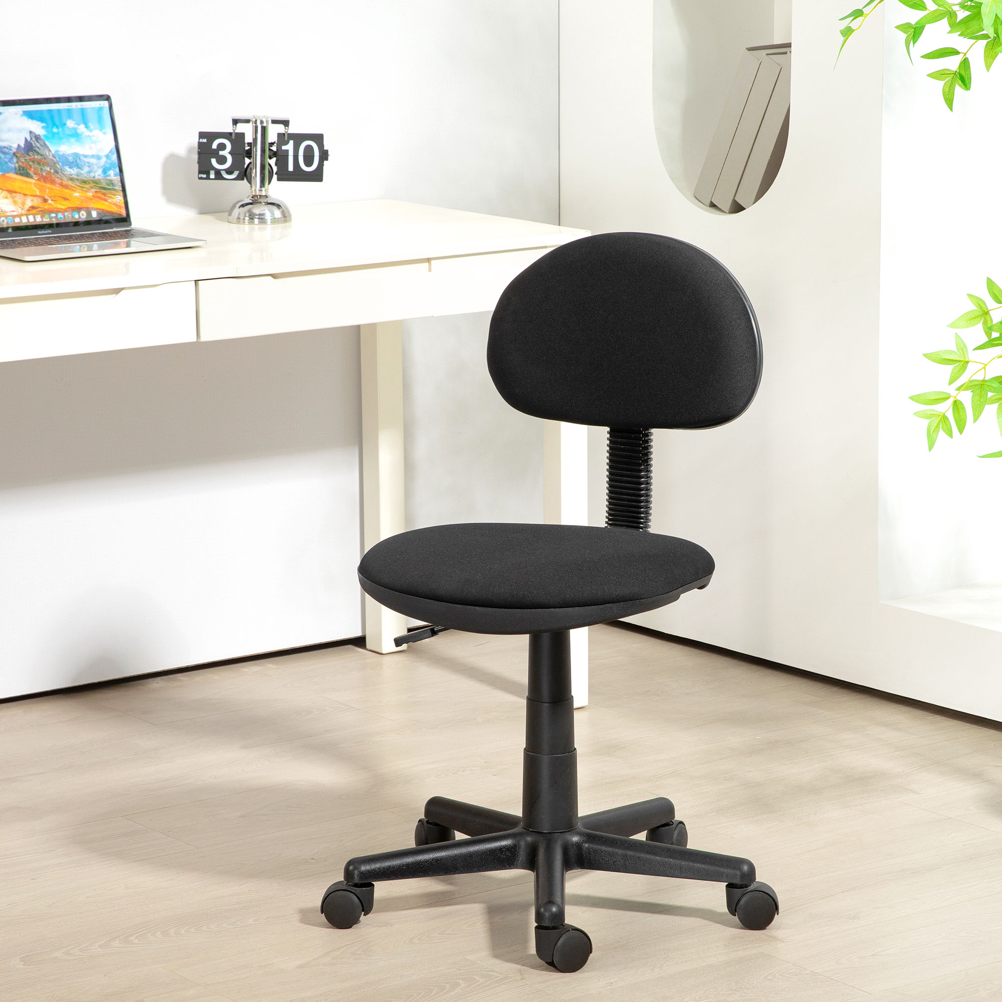 Una sedia da ufficio nera in una stanza luminosa. Una scrivania con laptop e orologio sullo sfondo. Foglie verdi a destra.