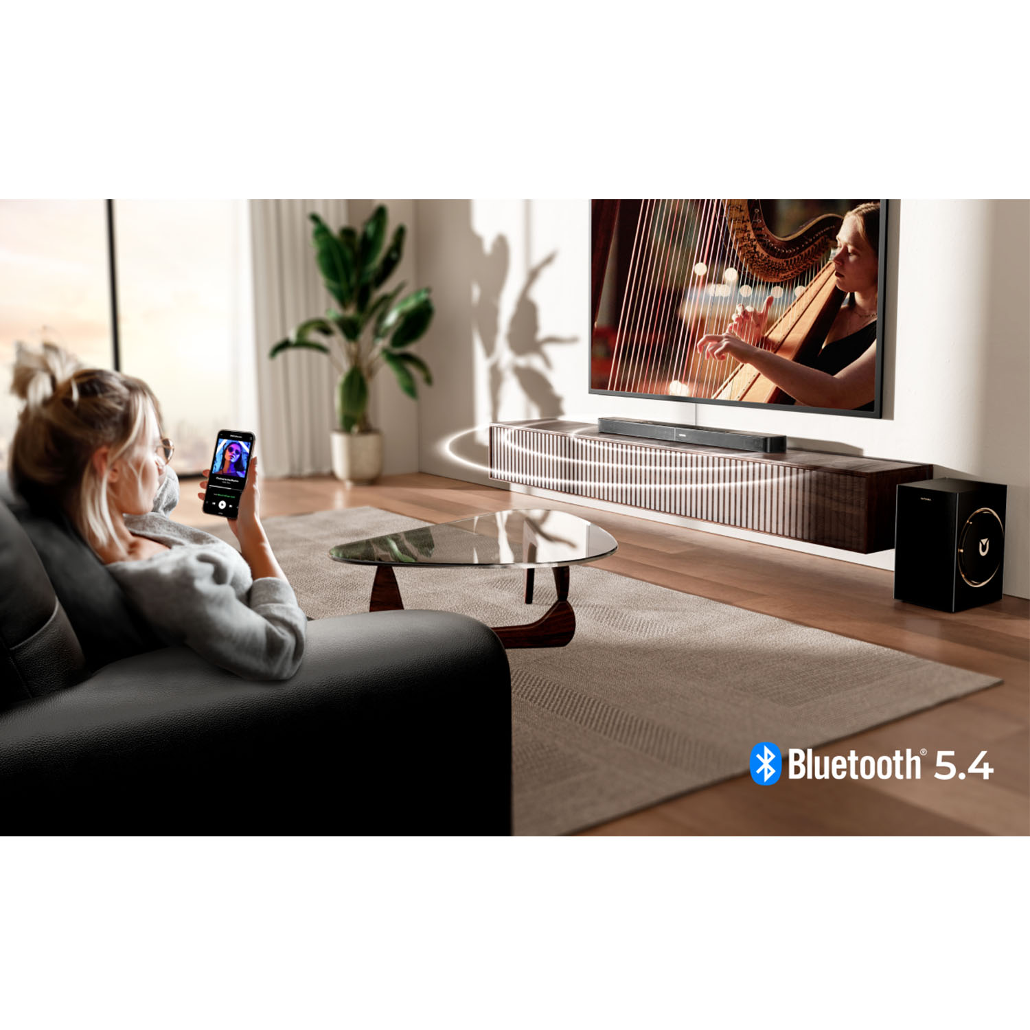 Kobieta siedzi na sofie, używając telefonu. Telewizor pokazuje harfistkę. Widoczny system dźwiękowy. Logo Bluetooth 5.4.
