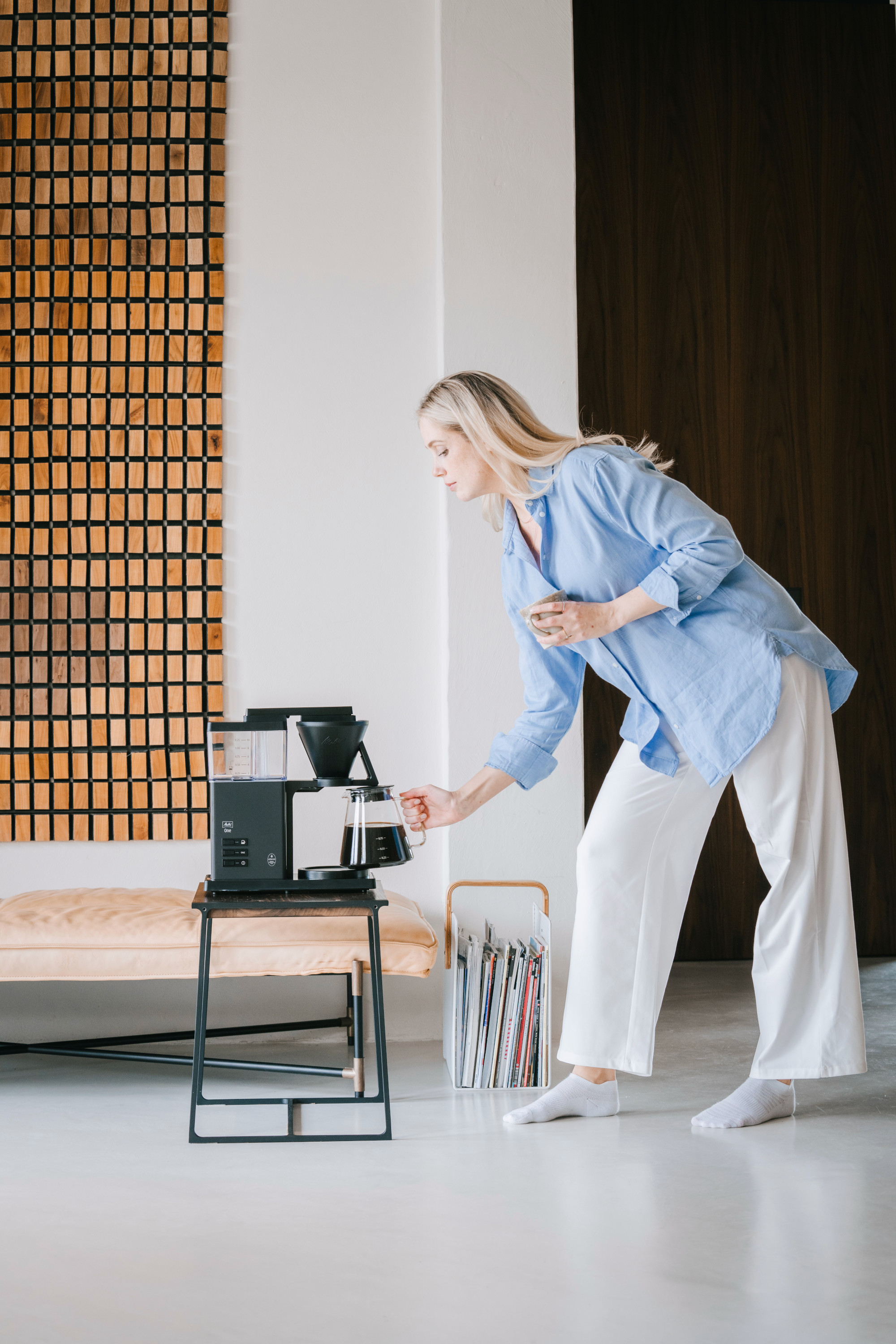 Kobieta w niebieskiej koszuli i białych spodniach nalewa kawę ze szklanej karafki. Ekspres do kawy i drewniane dzieło sztuki w tle.