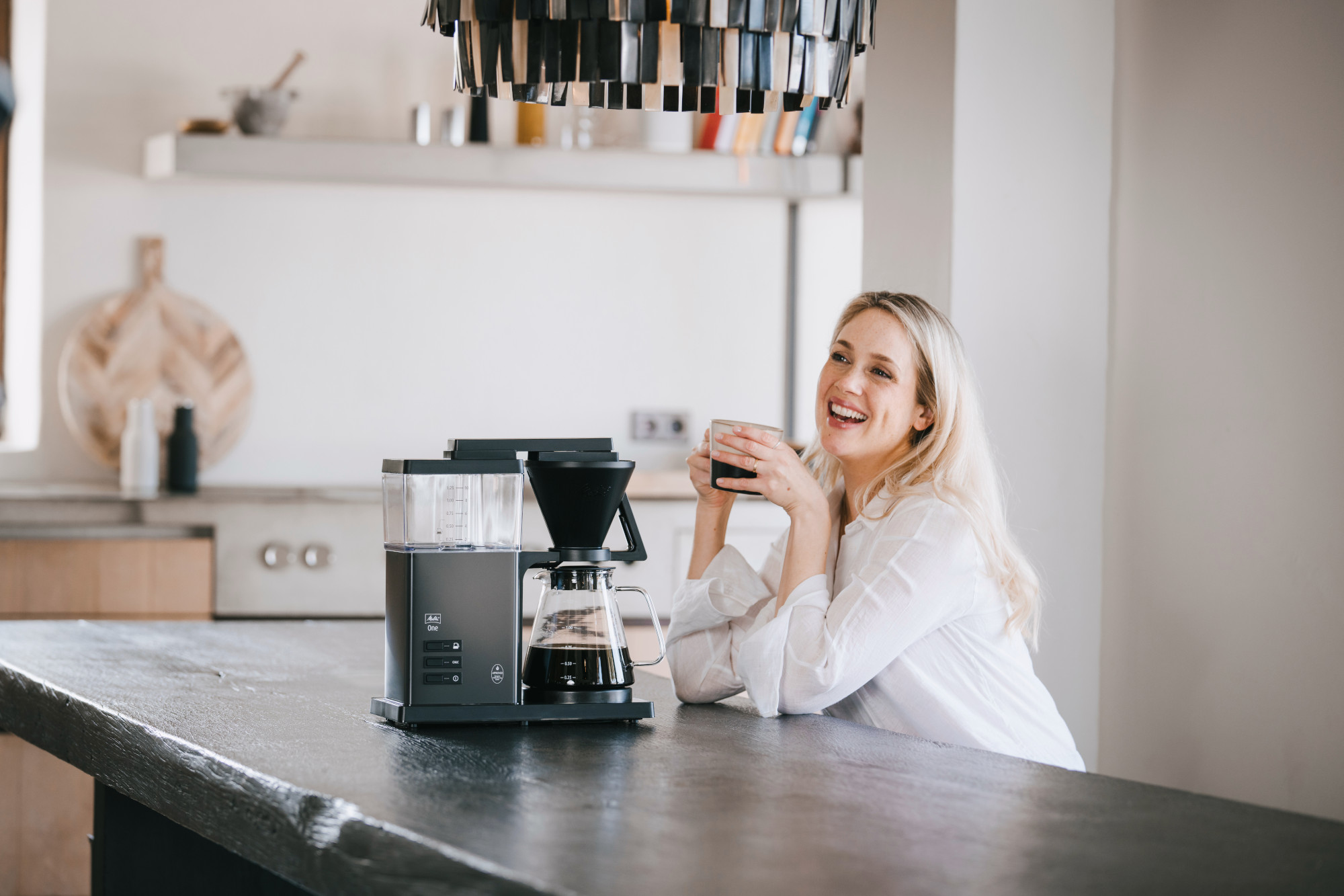 Kobieta w białej koszuli uśmiecha się, trzymając kubek w pobliżu ekspresu do kawy. W tle nowoczesna kuchnia.