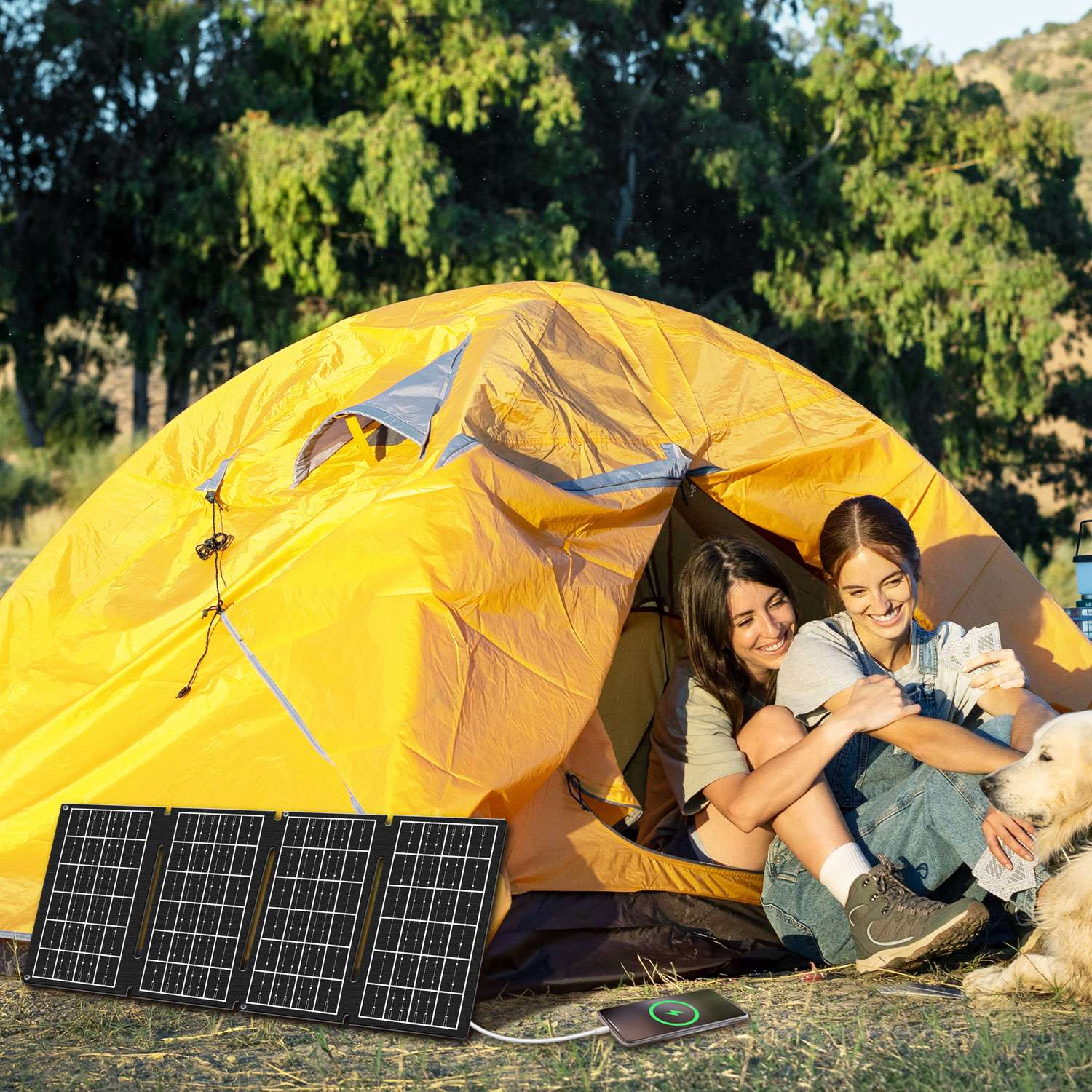 Dwie kobiety siedzą przed żółtym namiotem i grają w karty. Obok nich siedzi pies. Panele słoneczne ładują telefon. W tle drzewa.