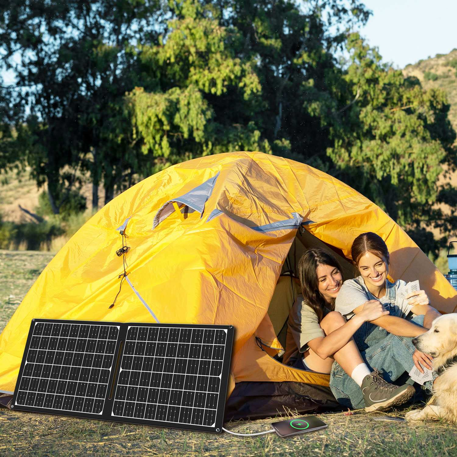 Due donne sedute in una tenda gialla con un golden retriever. Un pannello solare in primo piano, un telefono in carica nelle vicinanze.