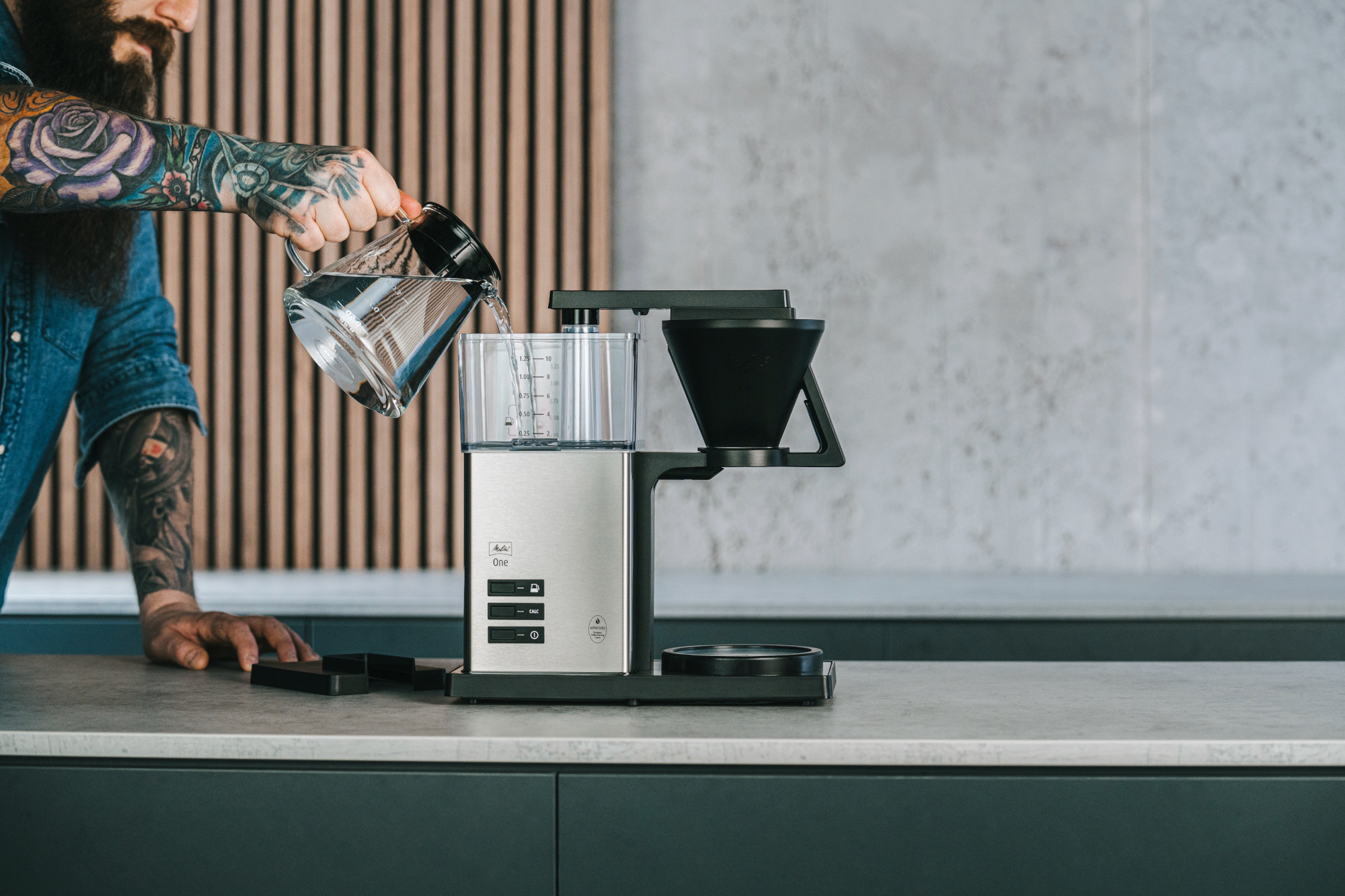 Un homme verse de l'eau d'une carafe en verre dans une cafetière. L'homme a des tatouages sur le bras. Le fond est moderne.