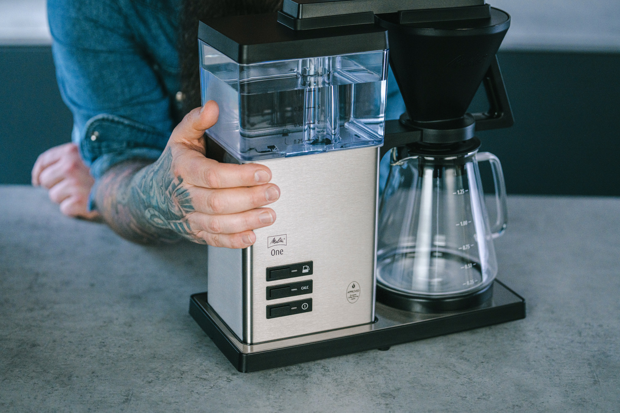 Une main tatouée tient une cafetière argentée. Une carafe en verre et un filtre noir reposent sur une base en métal.