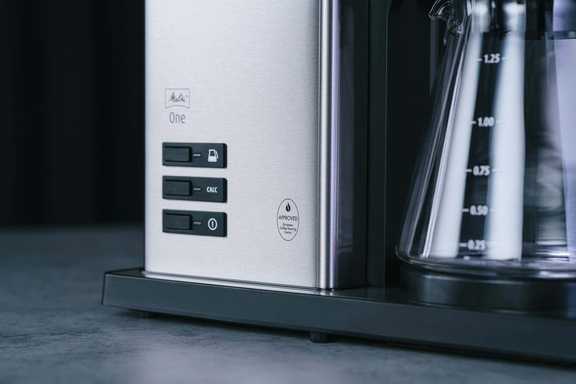 Gros plan sur une cafetière. Corps en acier inoxydable avec boutons. Une carafe en verre avec des marquages. La cafetière est sur une surface sombre.
