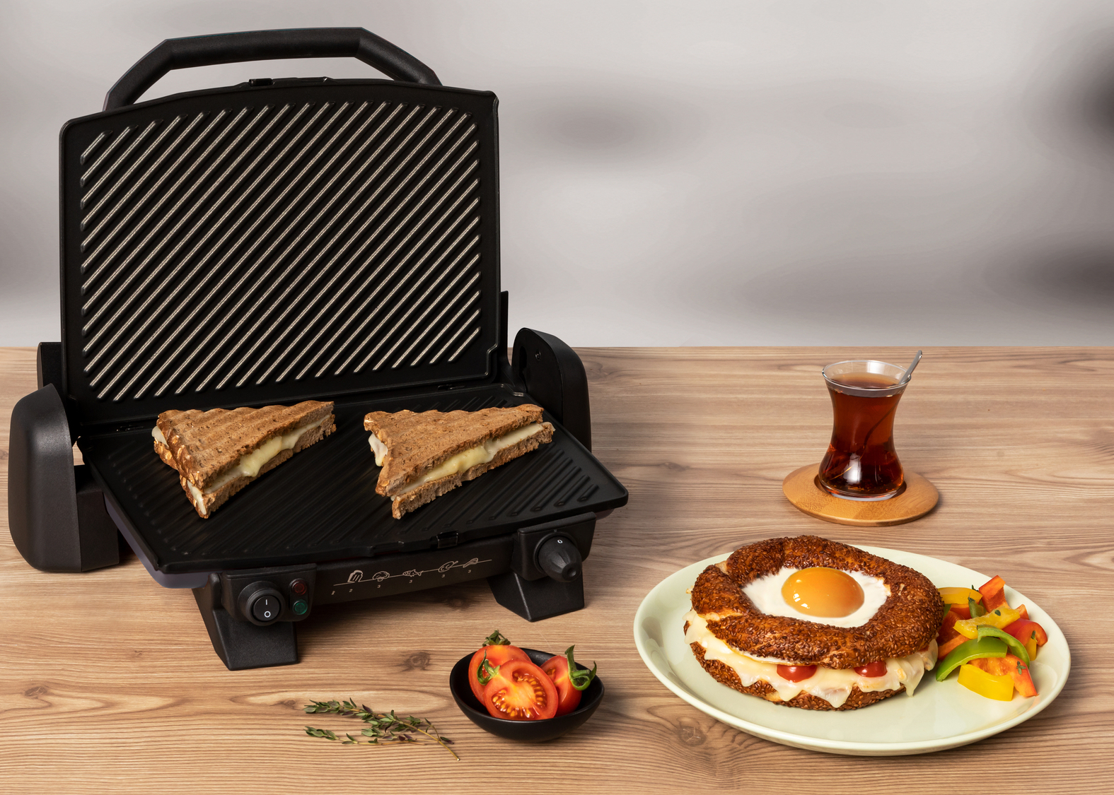 Izgara sandviç makinesi, yumurtalı simit, domates, biberler ve ahşap bir masada bir bardak çay.