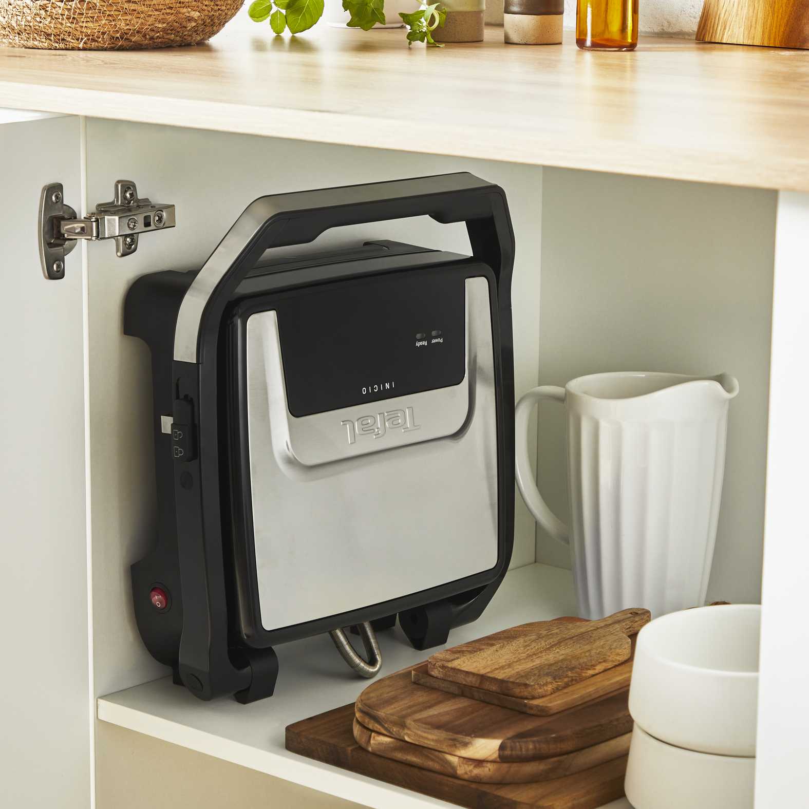Czarny i srebrny opiekacz do kanapek jest w białej szafce. Drewniane deski do krojenia i biały dzbanek są na półce.