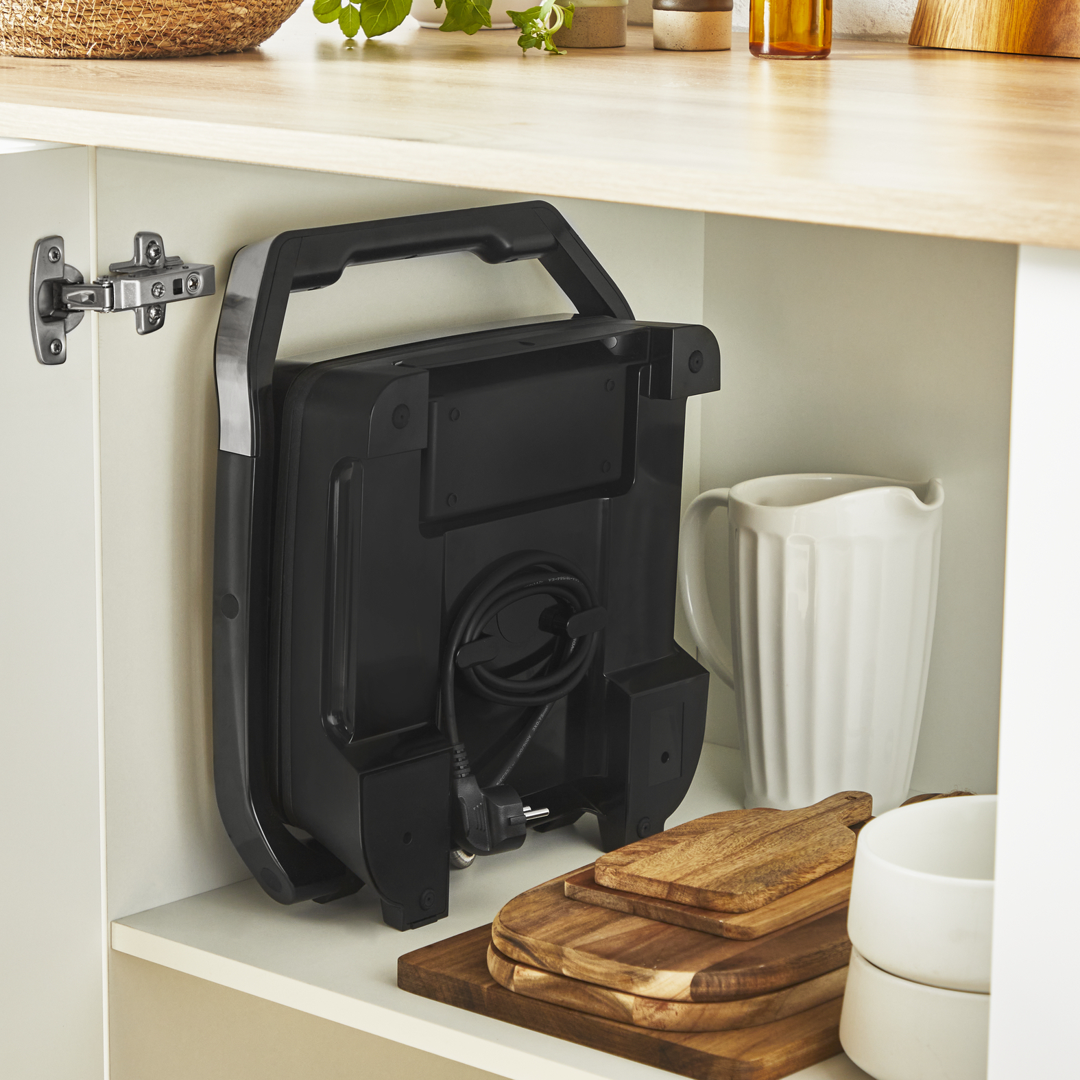 Czarny opiekacz do kanapek przechowywany w szafce kuchennej. Widoczny jest również biały dzbanek i drewniane deski do krojenia.