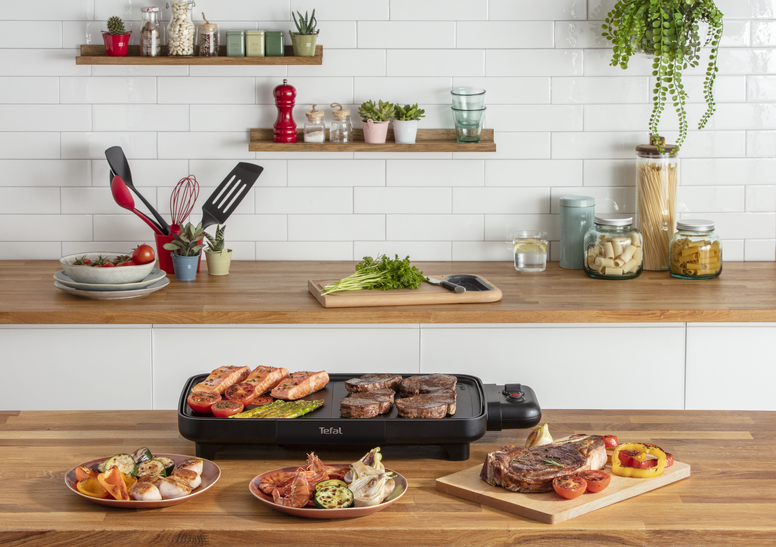 Una escena de cocina con una parrilla y comida cocinada: bistec, salmón, verduras. Fondo: azulejos blancos, estantes y encimera de madera.