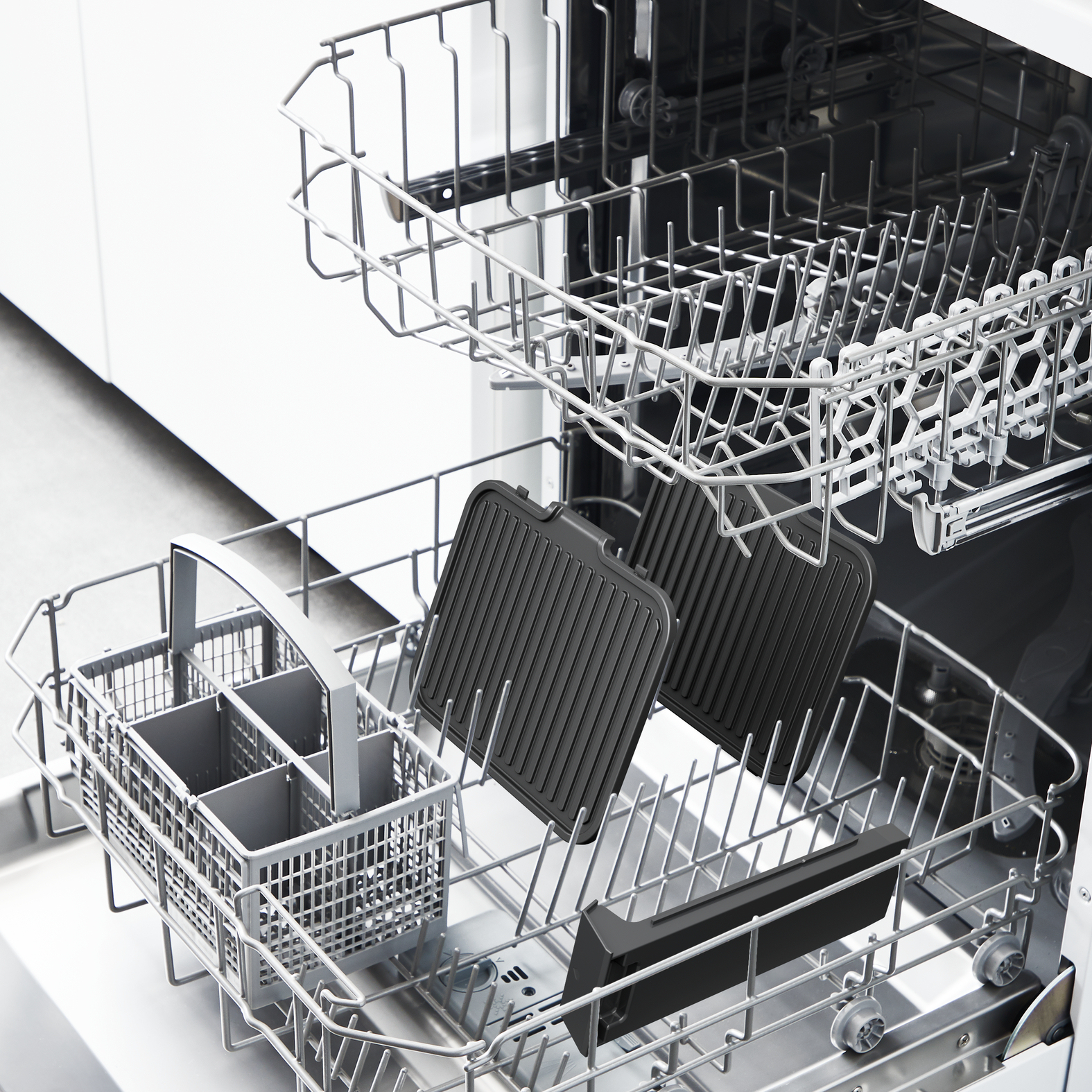 Intérieur de lave-vaisselle avec deux plaques de grill noires, un panier à couverts et une poignée noire. Des grilles argentées sont visibles.