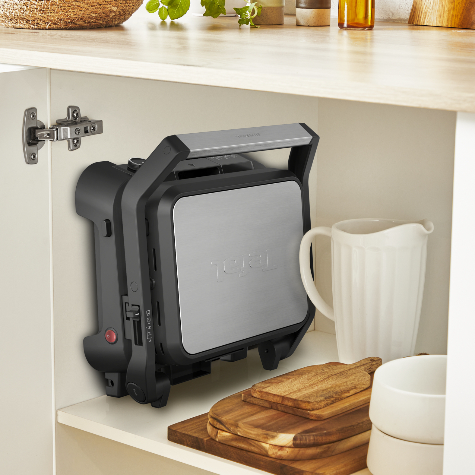 Un Supergrill Tefal est dans un placard blanc. Un pichet blanc et des planches en bois sont à proximité. Le grill est noir et argenté.