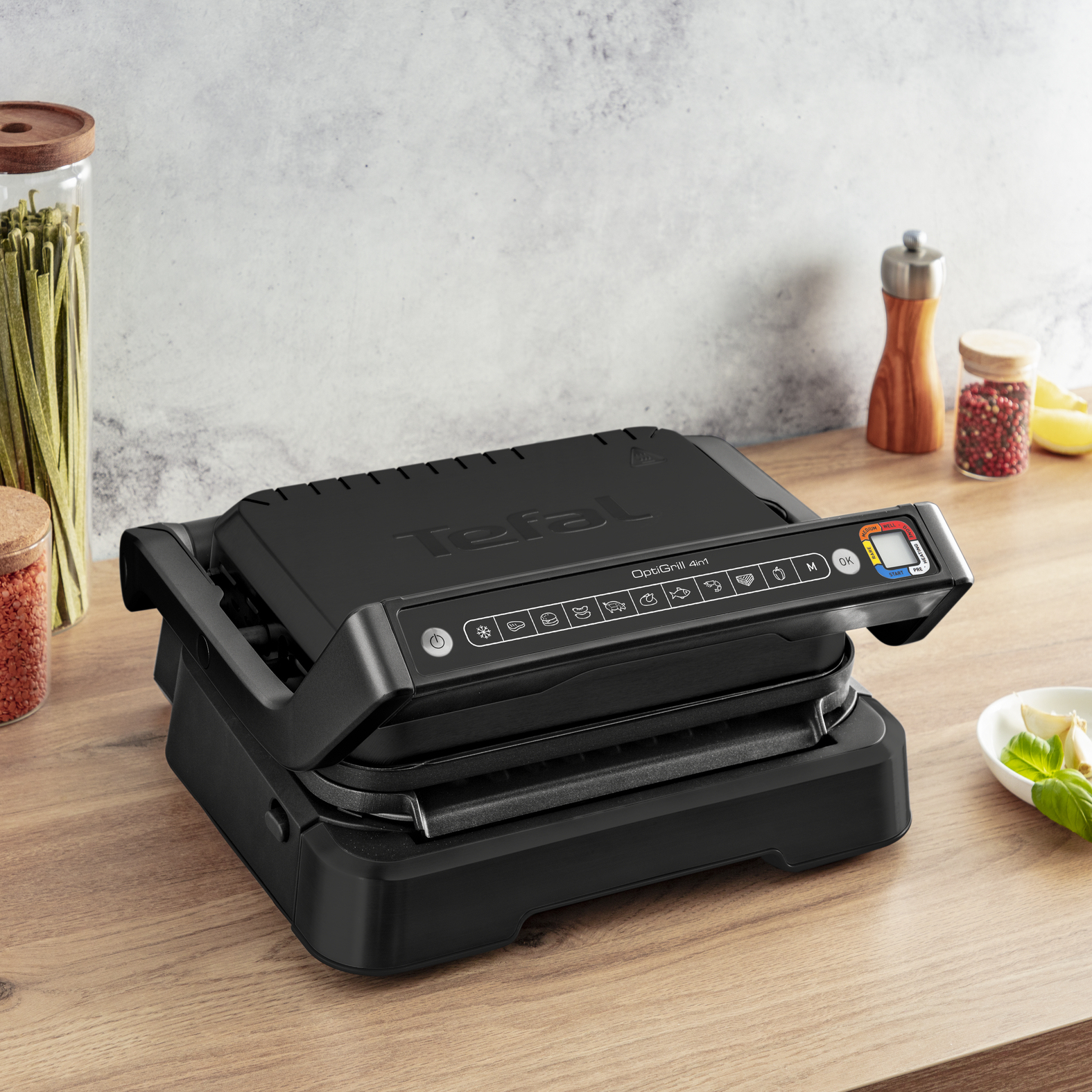 Un Tefal OptiGrill 4in1 nero su una superficie di legno. Sullo sfondo barattoli con ingredienti e un macinapepe.