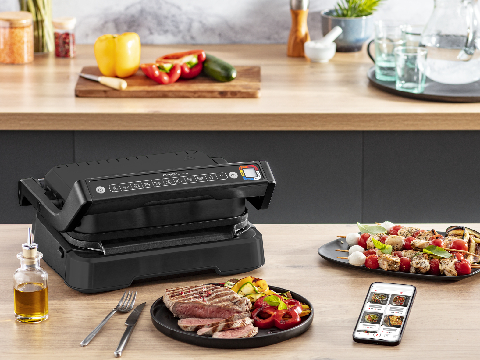 Un OptiGrill 4in1 nero con bistecca cotta, spiedini e verdure su un tavolo di legno, con un telefono che mostra una ricetta.