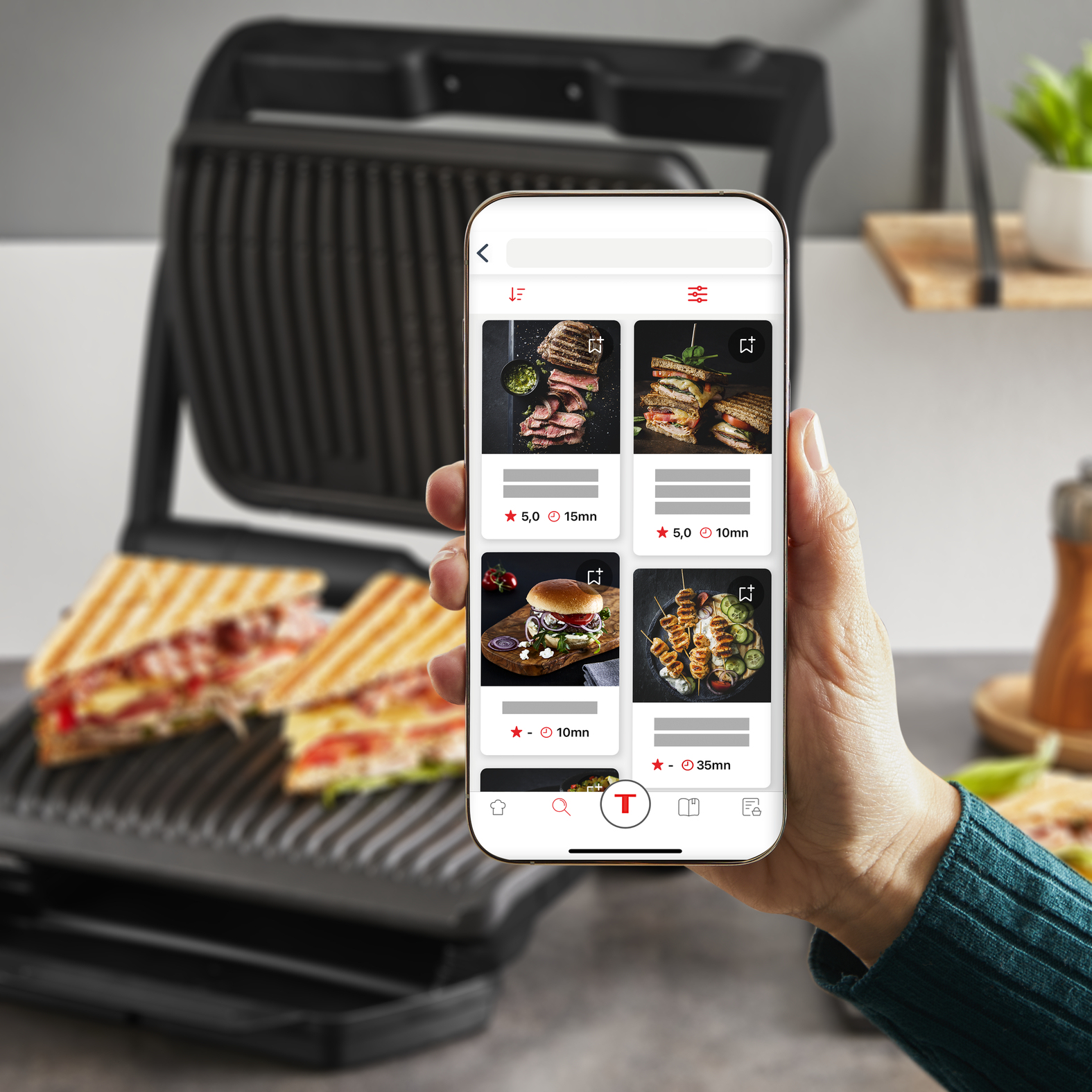 Ręka trzyma telefon z przepisami. Grill z kanapkami w tle.