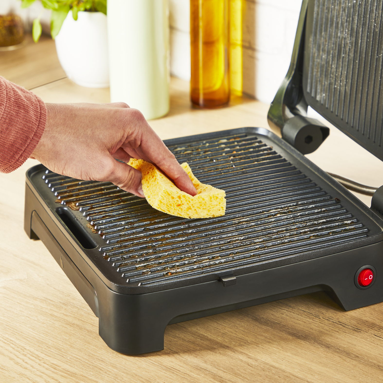 Ręka czyści brudny czarny grill żółtą gąbką. Grill stoi na drewnianej powierzchni, z czerwonym przyciskiem.