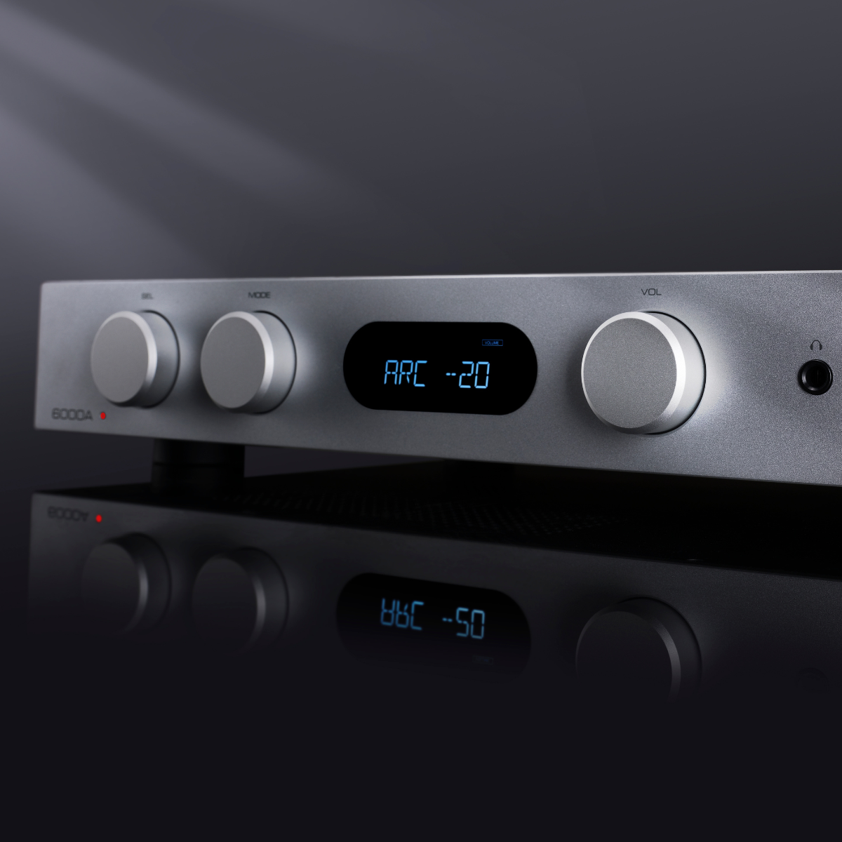 A silver audio amplifier with knobs, a display showing 'ARC -20', and a headphone jack. Reflected below on a dark surface.