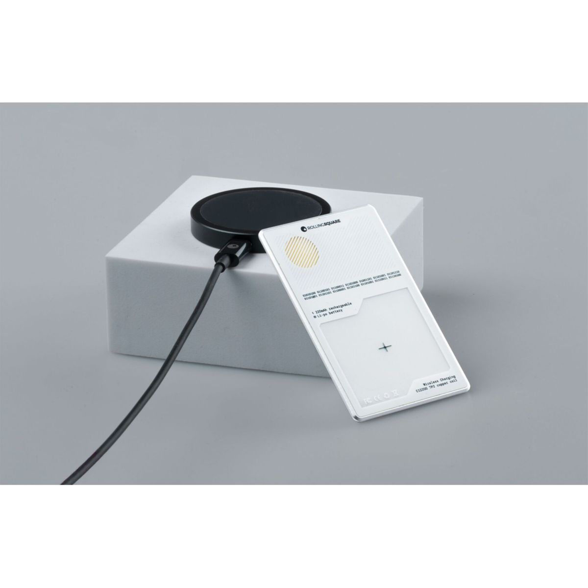 A black wireless charger and a white card are on a white block. A black cable connects the charger. The background is gray.
