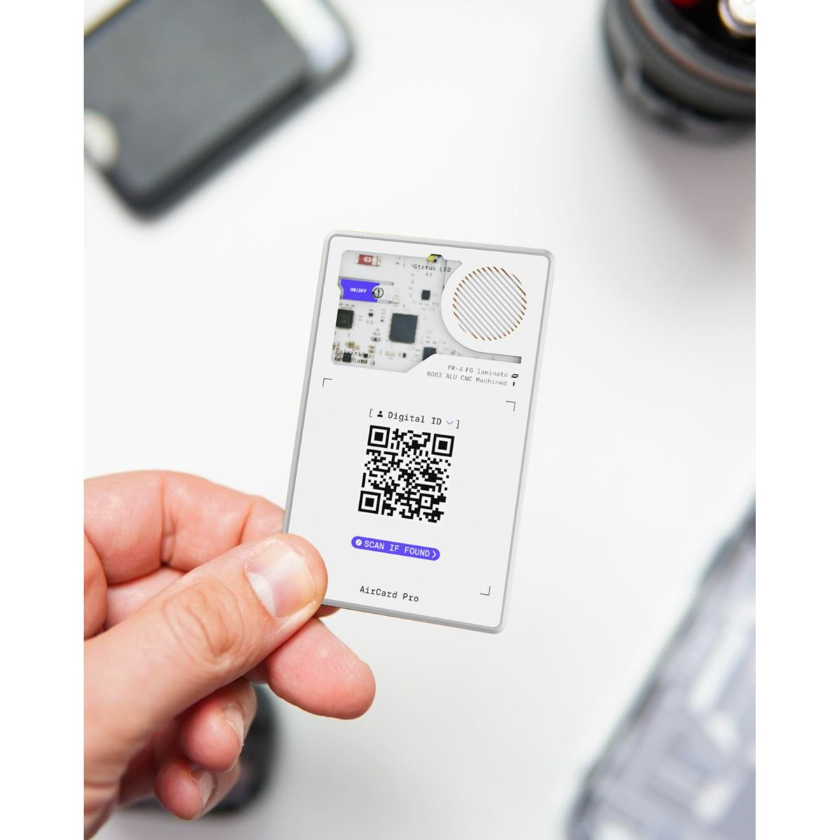 Hand holding a white AirCard Pro with a QR code, a digital ID label, and 'Scan if Found' button. The background is blurred.