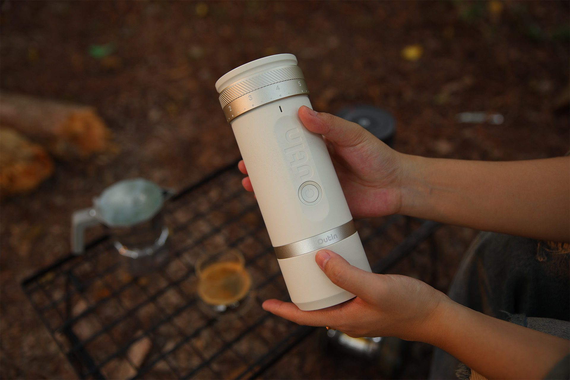 Osoba trzyma biało-srebrny przenośny ekspres do kawy. Inny sprzęt kempingowy w rozmytym tle.