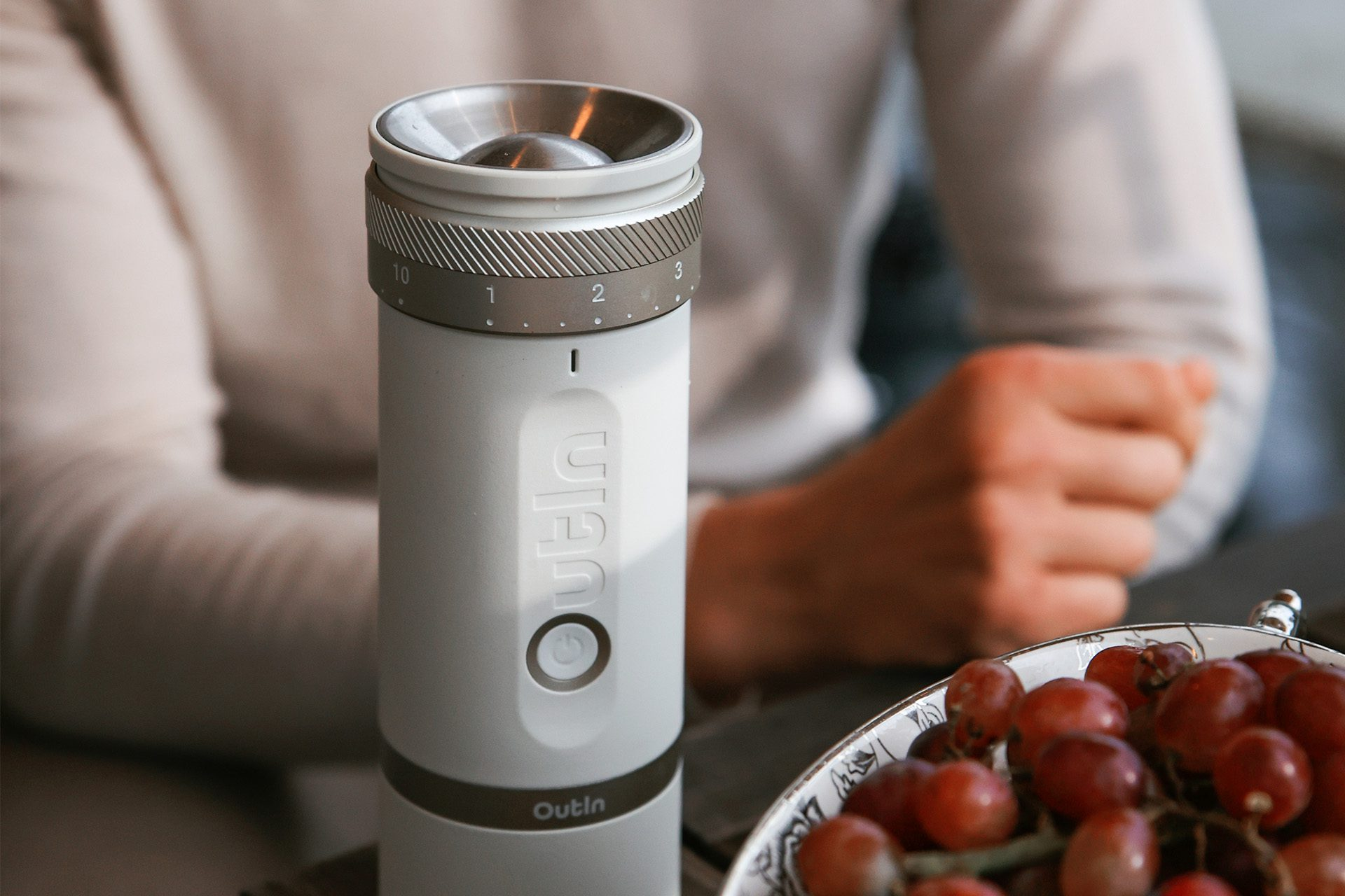 Biały młynek do kawy stoi na stole. Winogrona w misce na pierwszym planie, osoba w tle.